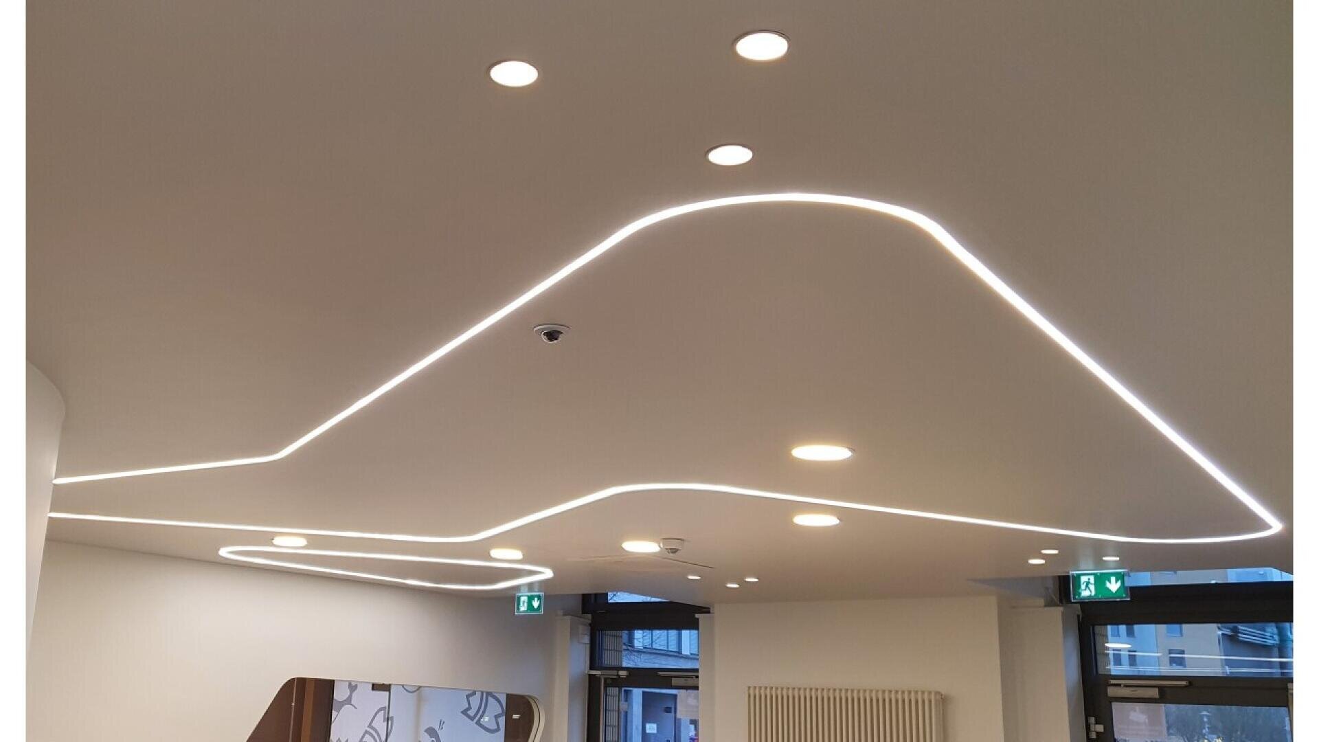 A modern white ceiling with embedded round spotlights and curved, continuous LED strip lighting; large windows and exit signs are visible along the walls below.