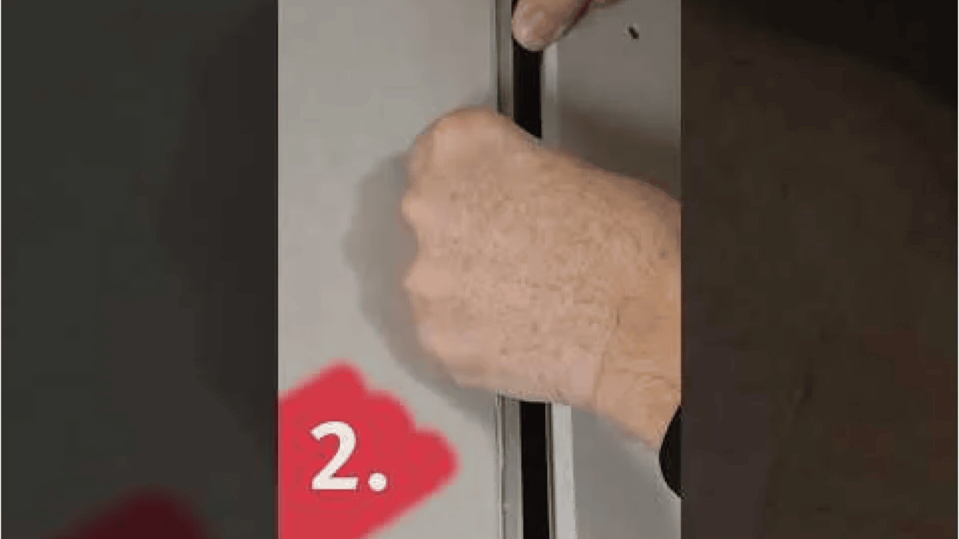 Eine Hand hebelt mit Gewalt die Naht zwischen zwei grauen Flächen auf, möglicherweise einer Tür oder einem Schrank. In der unteren linken Ecke ist eine rote Zahl 2 zu sehen.