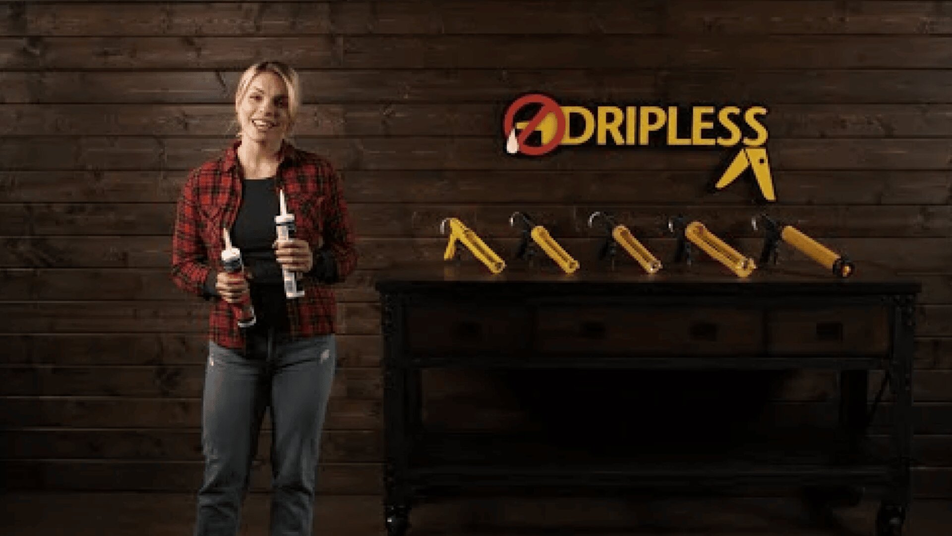A woman in a red plaid shirt holds two caulking guns, standing beside a table displaying more caulking guns. Behind her is a wooden wall with a DRIPLESS logo in yellow letters.