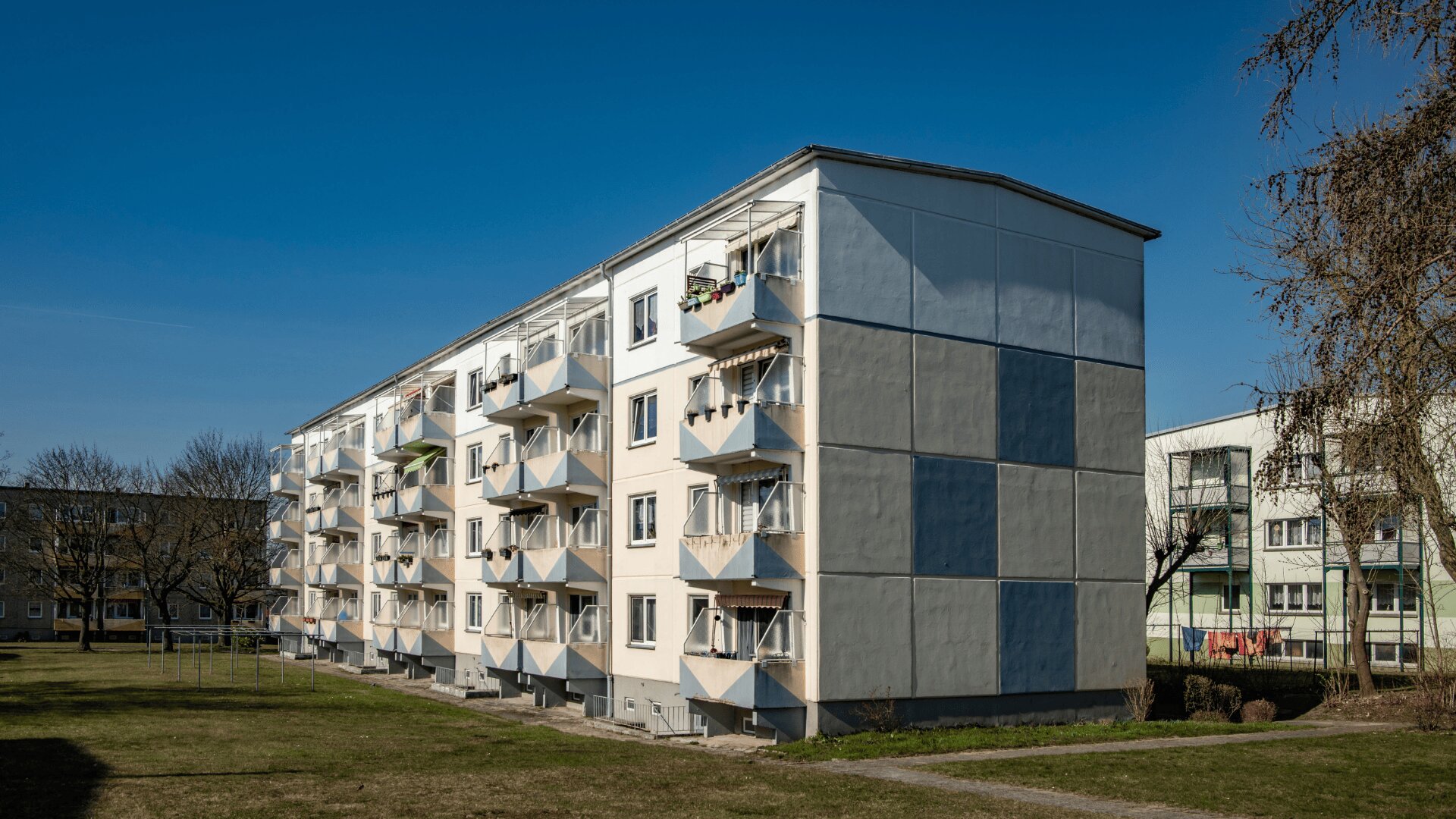 Ein vierstöckiges, weiß und grau gestrichenes Wohnhaus mit Balkonen steht auf einer Rasenfläche mit Bäumen unter einem strahlend blauen Himmel. Im Hintergrund sind weitere Wohnhäuser zu sehen.
