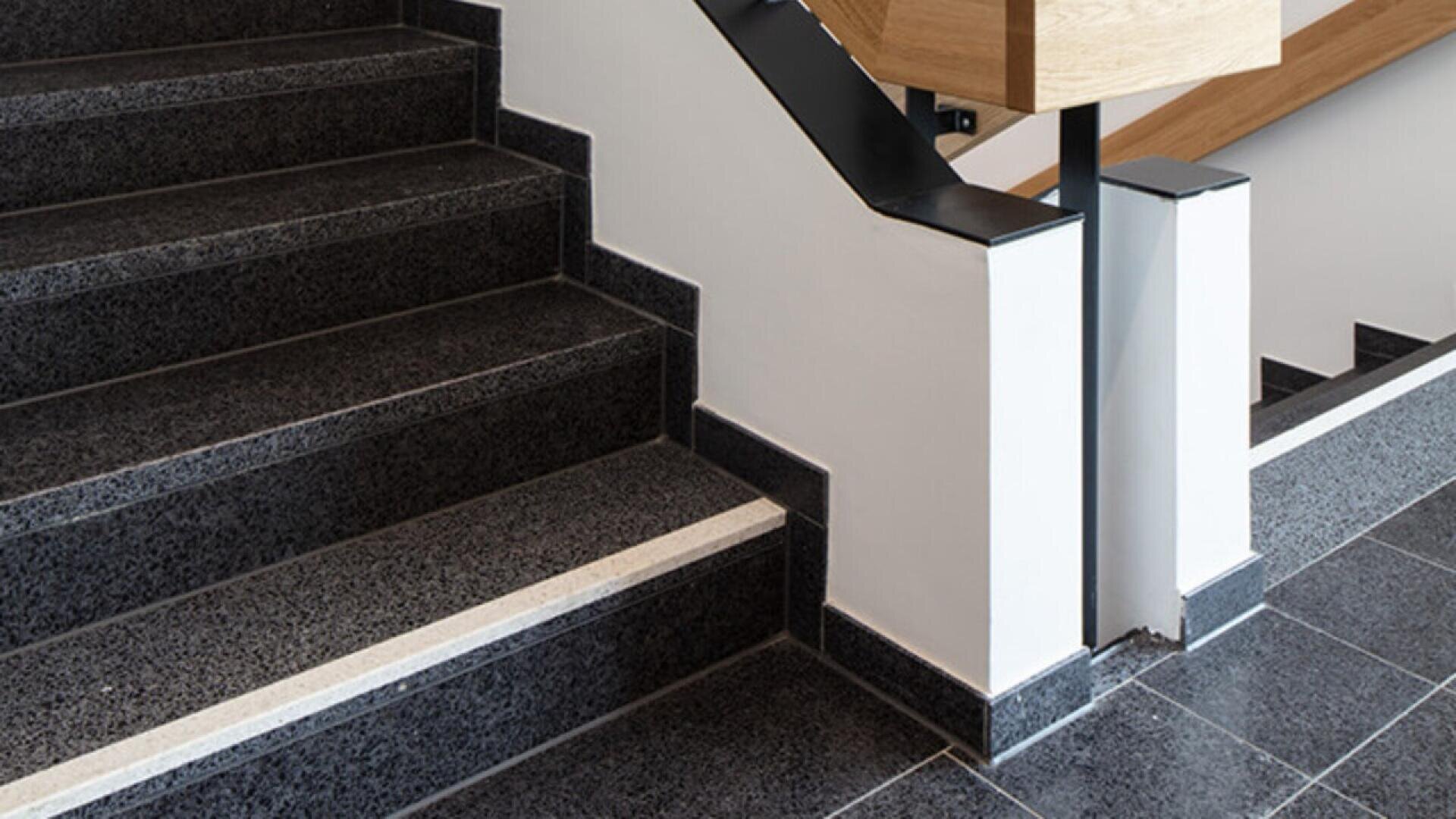 Black granite stairs with a white safety strip on the edge, next to a white wall and a modern wooden handrail with black metal brackets. The stairs have a clean, contemporary design.