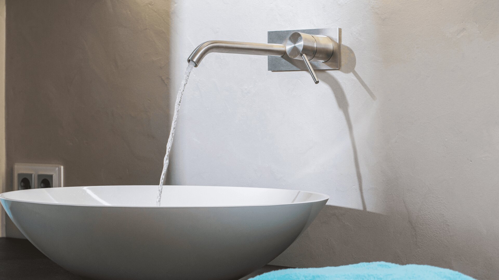 Ein moderner Wandhahn gießt Wasser in ein weißes, schüsselförmiges Waschbecken vor einer hellen, strukturierten Wand. In der unteren rechten Ecke ist ein blaues Handtuch zu sehen.