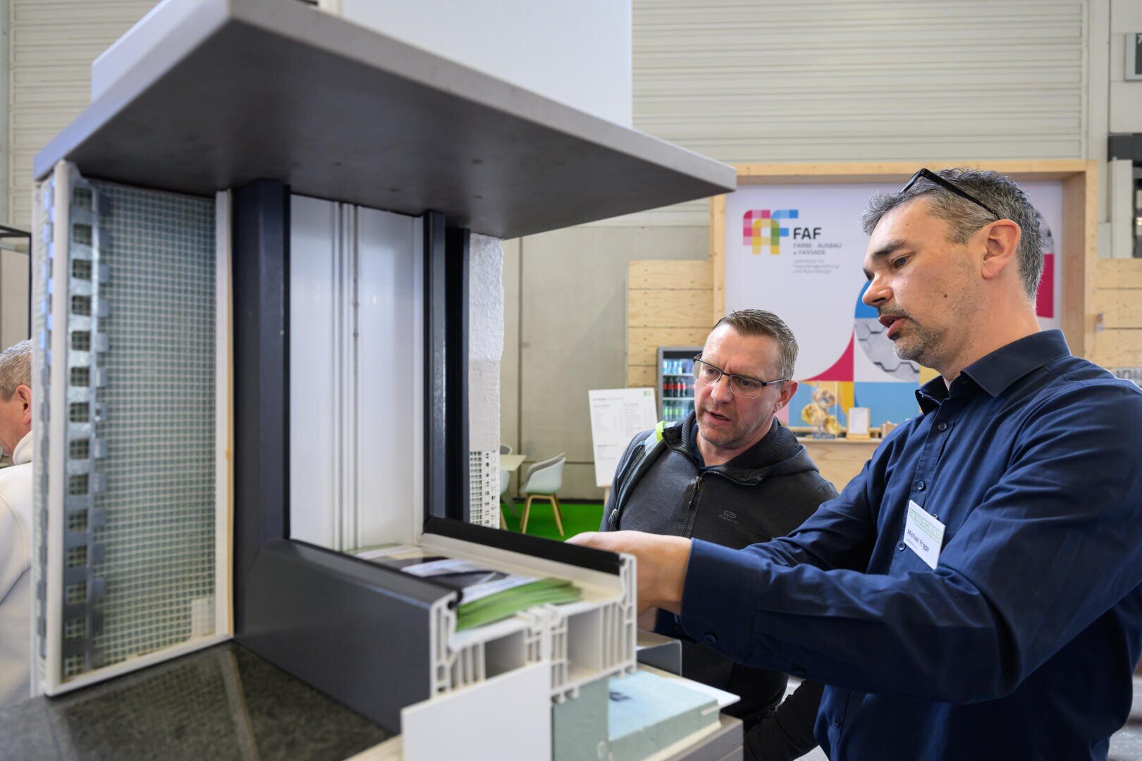 Zwei Männer betrachten und besprechen ein detailliertes Architekturmodell oder einen Gebäudeabschnitt auf einer Ausstellung oder Messe. Ein Mann zeigt auf einen Teil des Modells, während der andere aufmerksam zuhört.