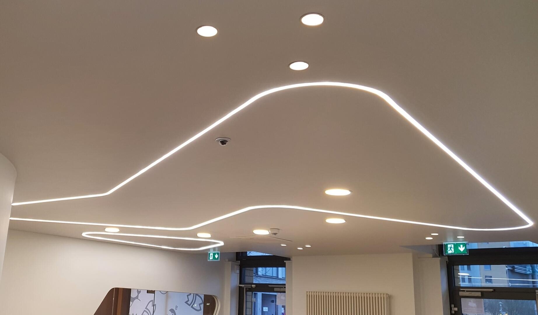 Moderne weiße Decke mit geschwungener, eingelassener LED-Lichtleiste und runden Strahlern. Zwei sichtbare Notausgangsschilder und Fenster schaffen einen hellen, modernen Innenraum.