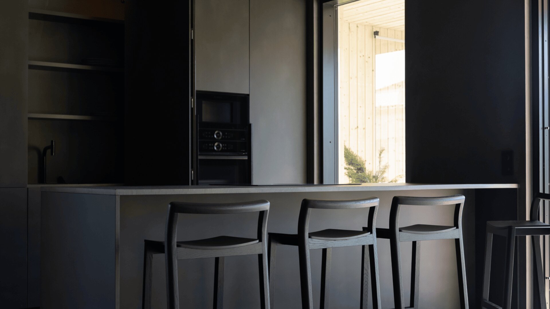 Eine moderne Küche mit drei Barhockern an einer minimalistischen Insel, dunklen Schränken und einem offenen Fenster, das natürliches Licht hereinlässt. Der Raum hat ein schlichtes, zeitgenössisches Design mit neutralen Farben.