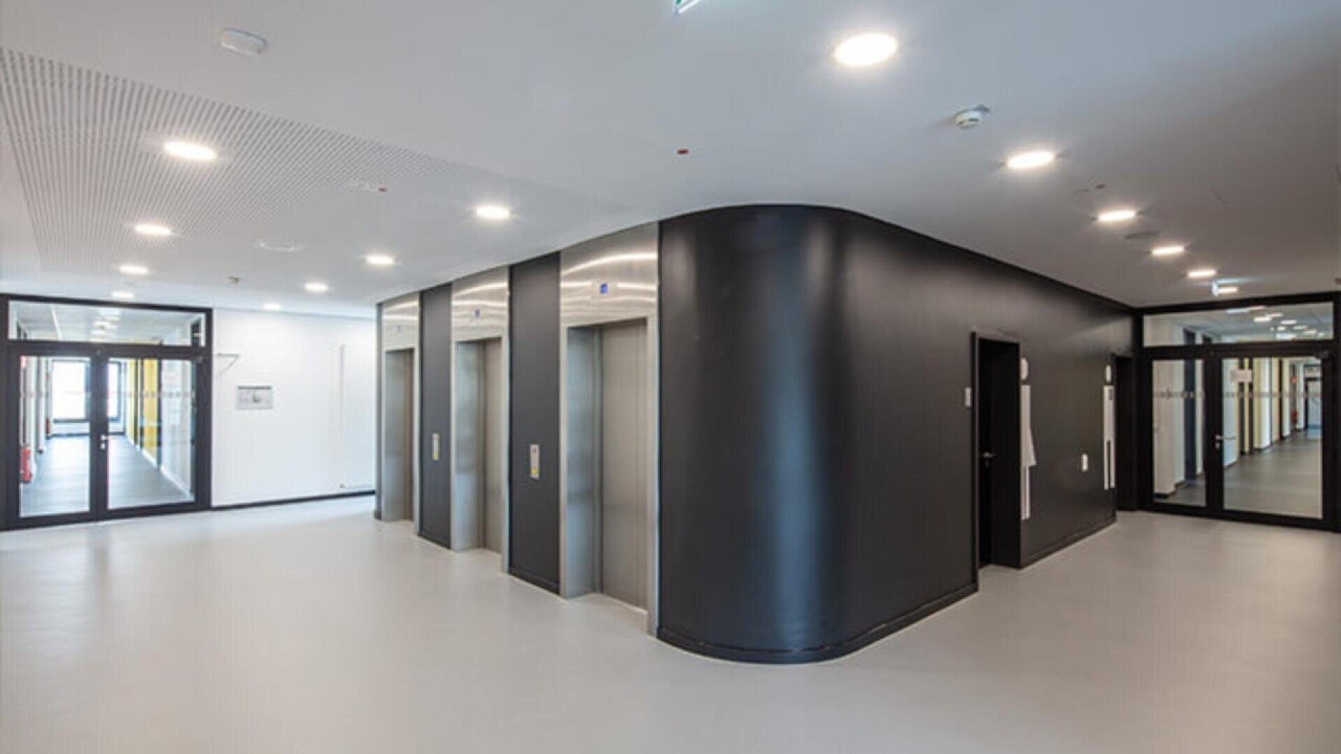 A modern building interior with three stainless steel elevators set in a curved black wall, bright lighting, white floors, and glass doors leading to adjacent hallways.