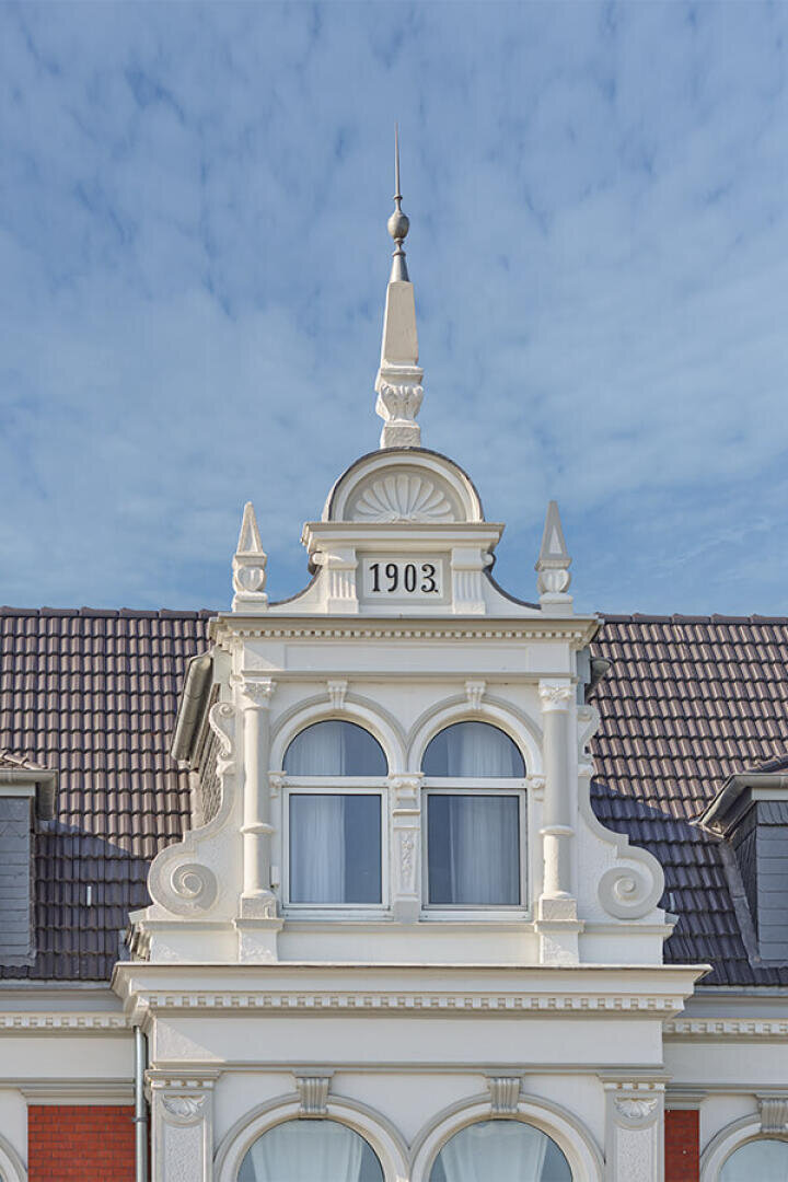 Verzierte historische Gebäudefassade mit dekorativen weißen Zierleisten, Bogenfenstern und einem Turm. Die Zahl 1903 steht in Dachnähe und weist auf das Baujahr hin. Der Himmel ist klar, mit leichten Wolken.