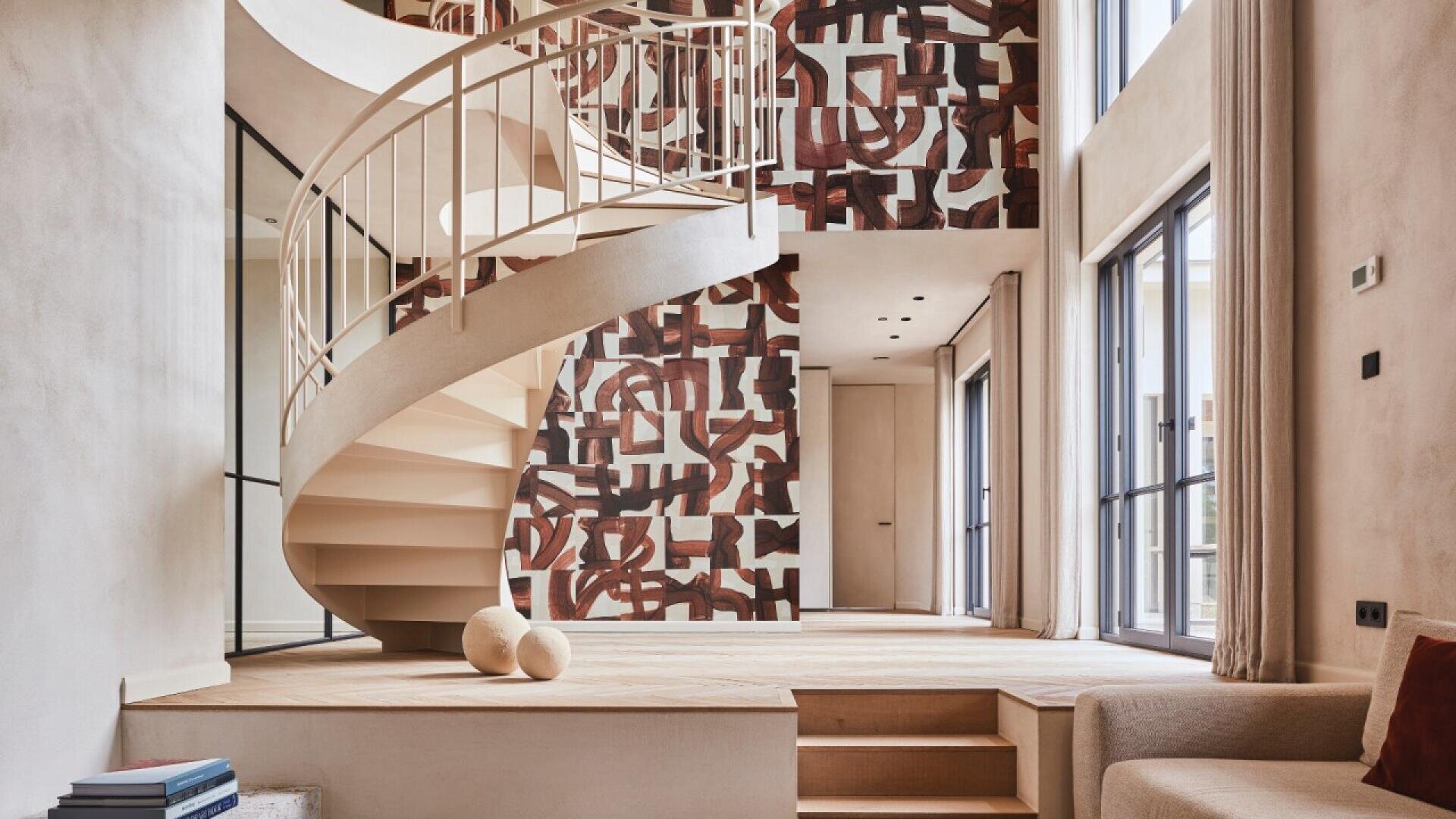 A modern living space with a white spiral staircase, abstract geometric mural on the back wall, large windows, beige walls, and minimalistic decor including a light sofa and two spherical objects on the floor.