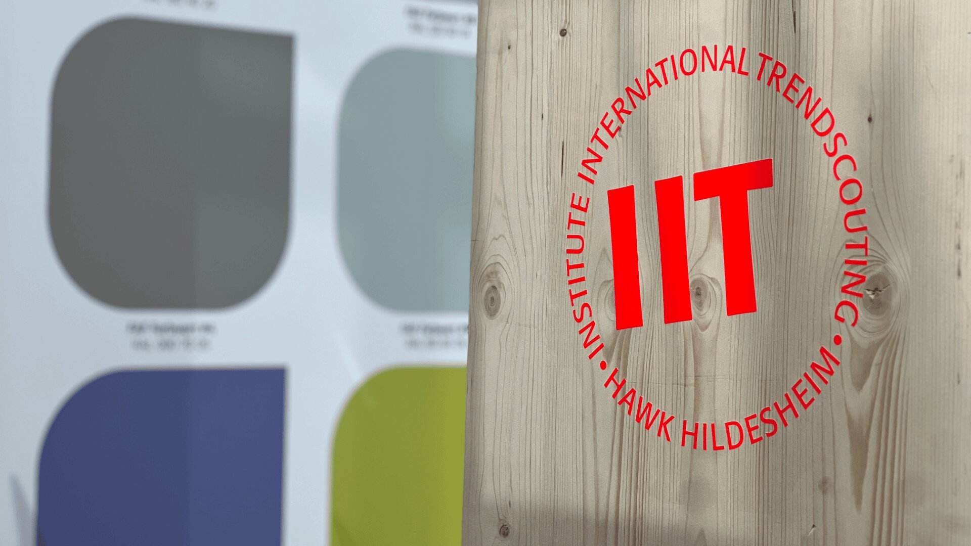 Eine Holztafel mit einem roten, runden Logo mit der Aufschrift „INSTITUTE INTERNATIONAL TRENDSCOUTING HAWK HILDESHEIM IIT“. Im Hintergrund befinden sich vier große farbige Quadrate mit Text darunter.