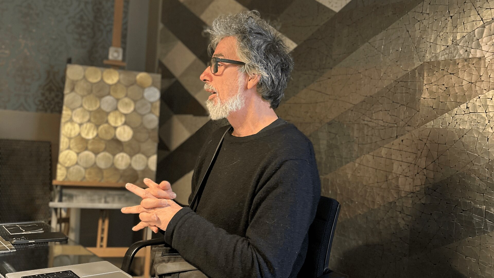 A man with gray hair and a beard, wearing glasses and a black sweater, sits at a desk with his hands clasped, facing left. Behind him are geometric-patterned walls and a board with circular shapes.