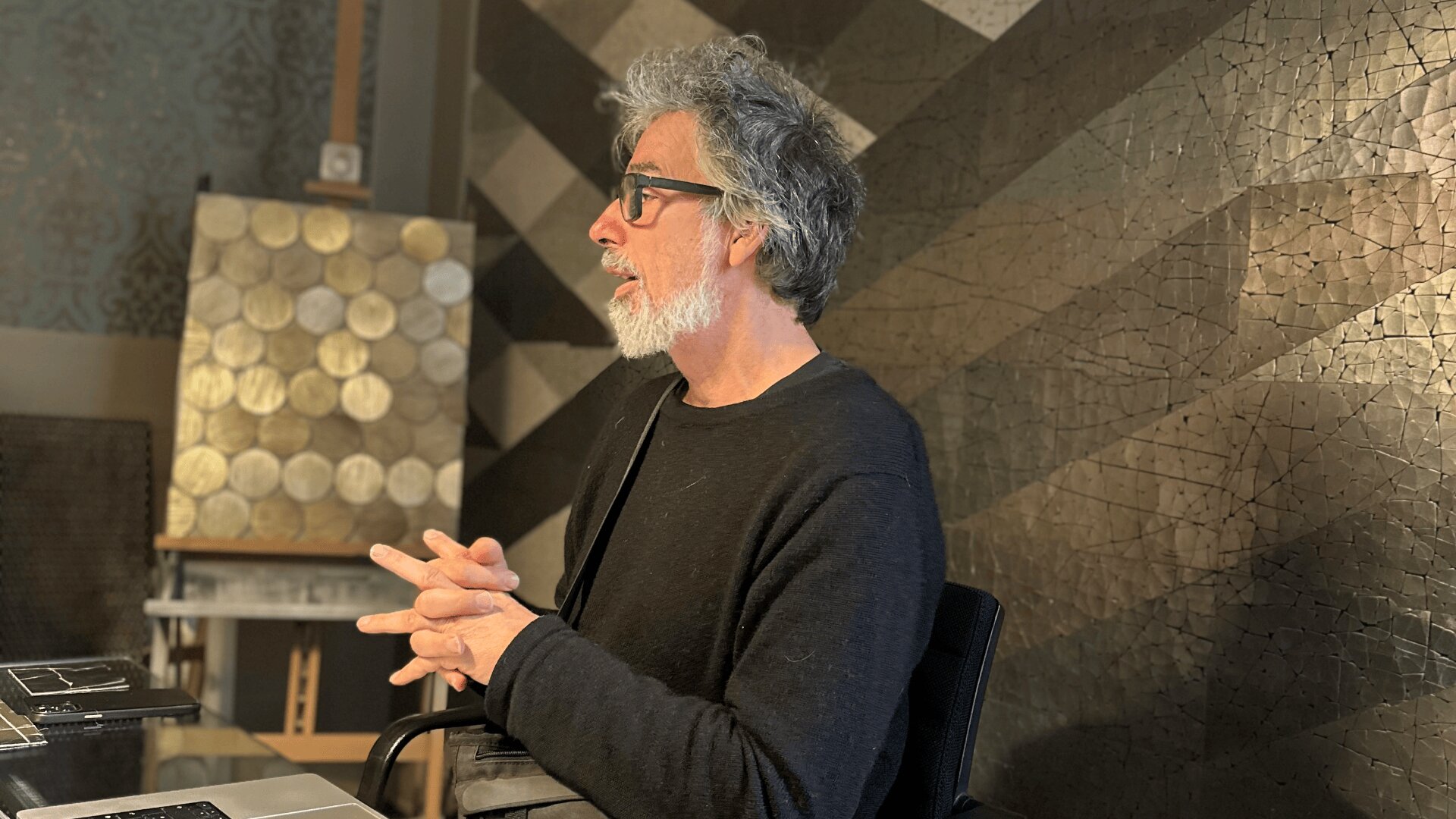 Ein Mann mit grauem Haar und Bart, Brille und dunklem Pullover sitzt mit gefalteten Händen an einem Schreibtisch und blickt nach links. Im Hintergrund sind geometrische Wandkunst und ein Panel mit kreisförmigen Mustern zu sehen.