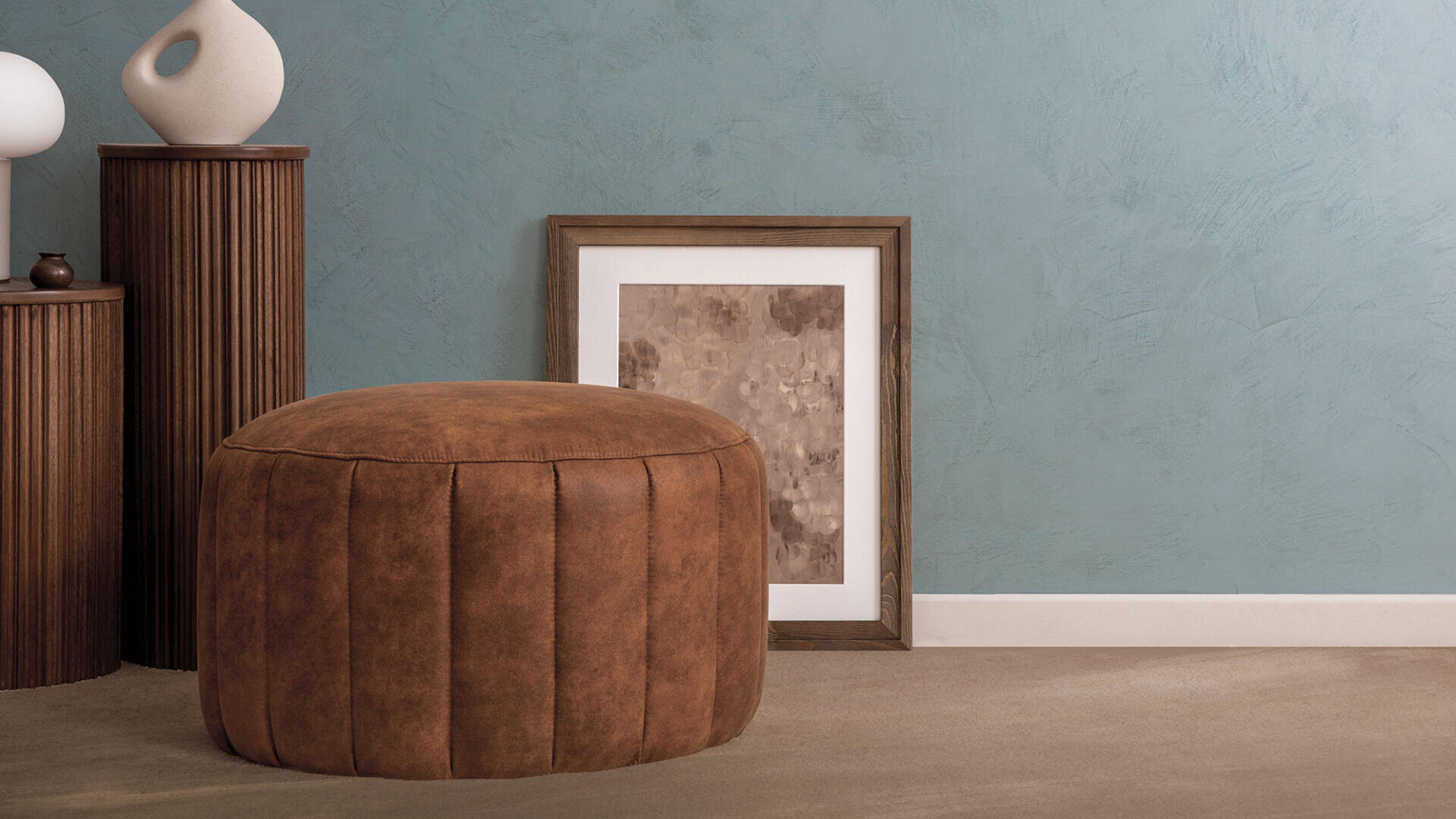 A brown leather ottoman sits on a beige carpet next to a textured blue wall. Beside it are wooden pedestals with decorative vases and a framed abstract artwork leaning against the wall.