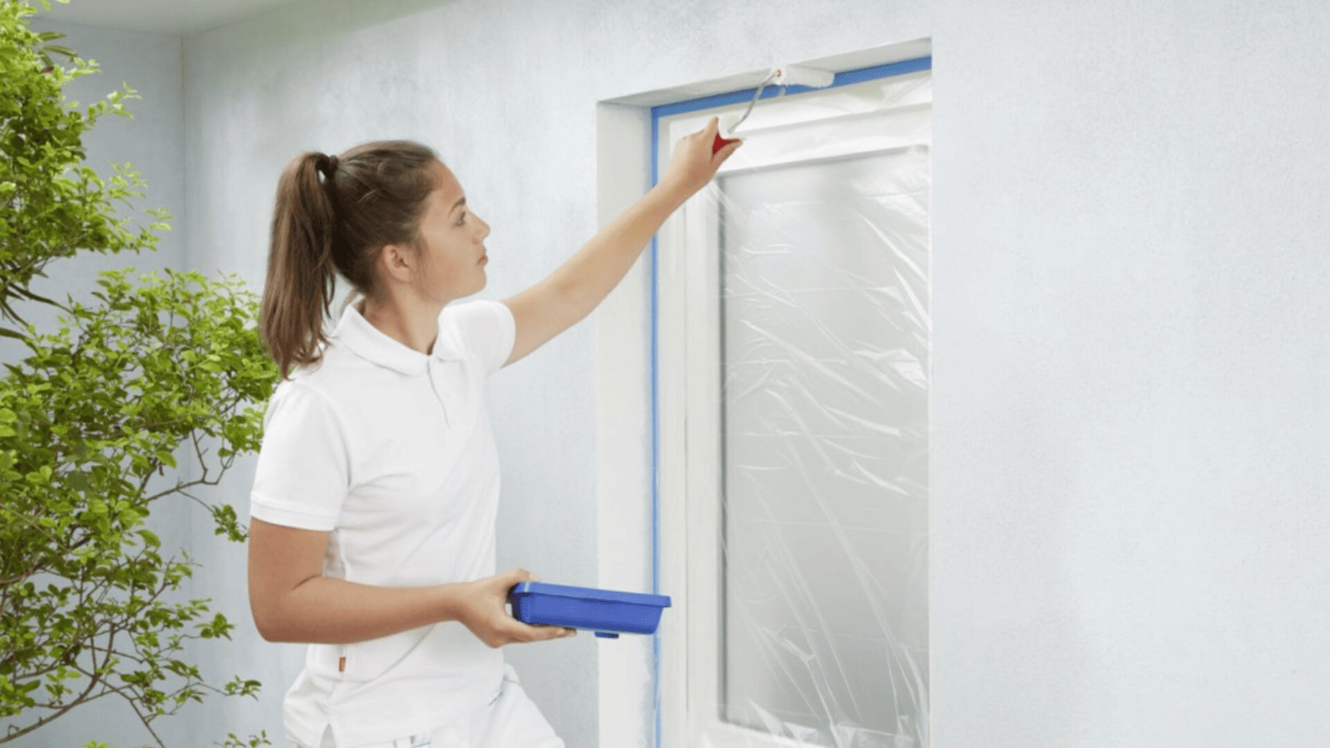 Eine Person in weißer Kleidung streicht mit einer Farbrolle die Außenwand um ein Fenster herum, das zum Schutz mit Plastikfolie und blauem Klebeband abgedeckt ist. In der Nähe ist eine grüne Blattpflanze zu sehen.
