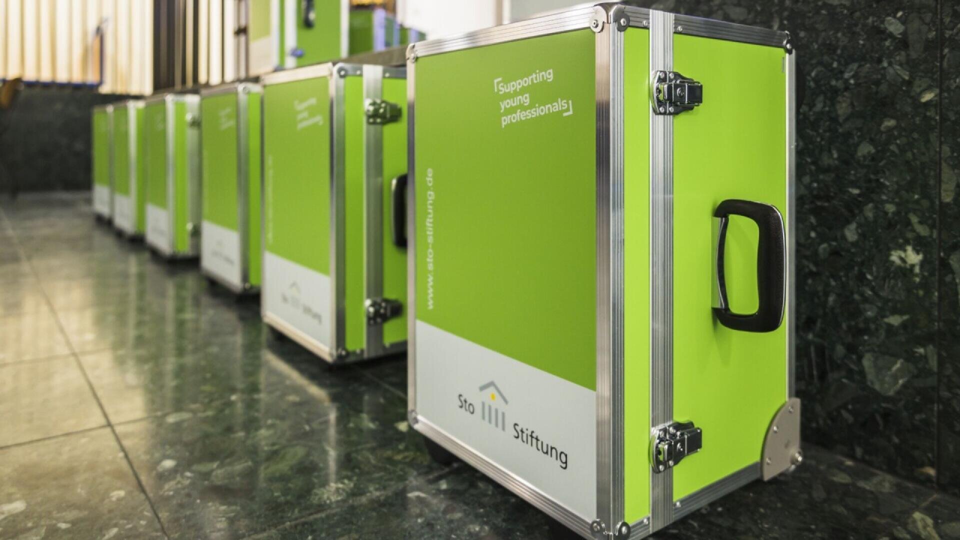 Several green and white boxes with black handles and metal edges are lined up on a shiny tile floor. The boxes display the logo and name Stiftung and the phrase Supporting young professionals.
