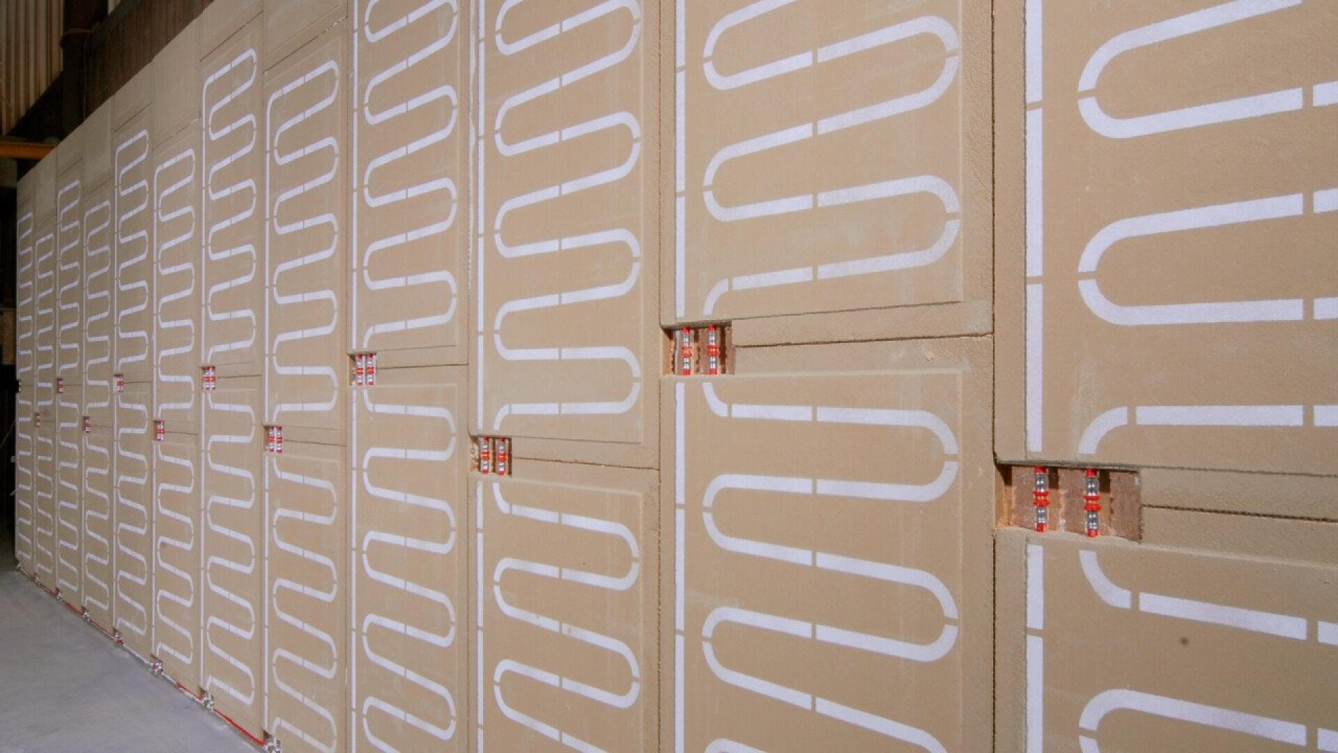 Large beige wall panels with white winding pipe patterns are stacked vertically in rows inside a building. Small orange connectors are visible at gaps between some panels.