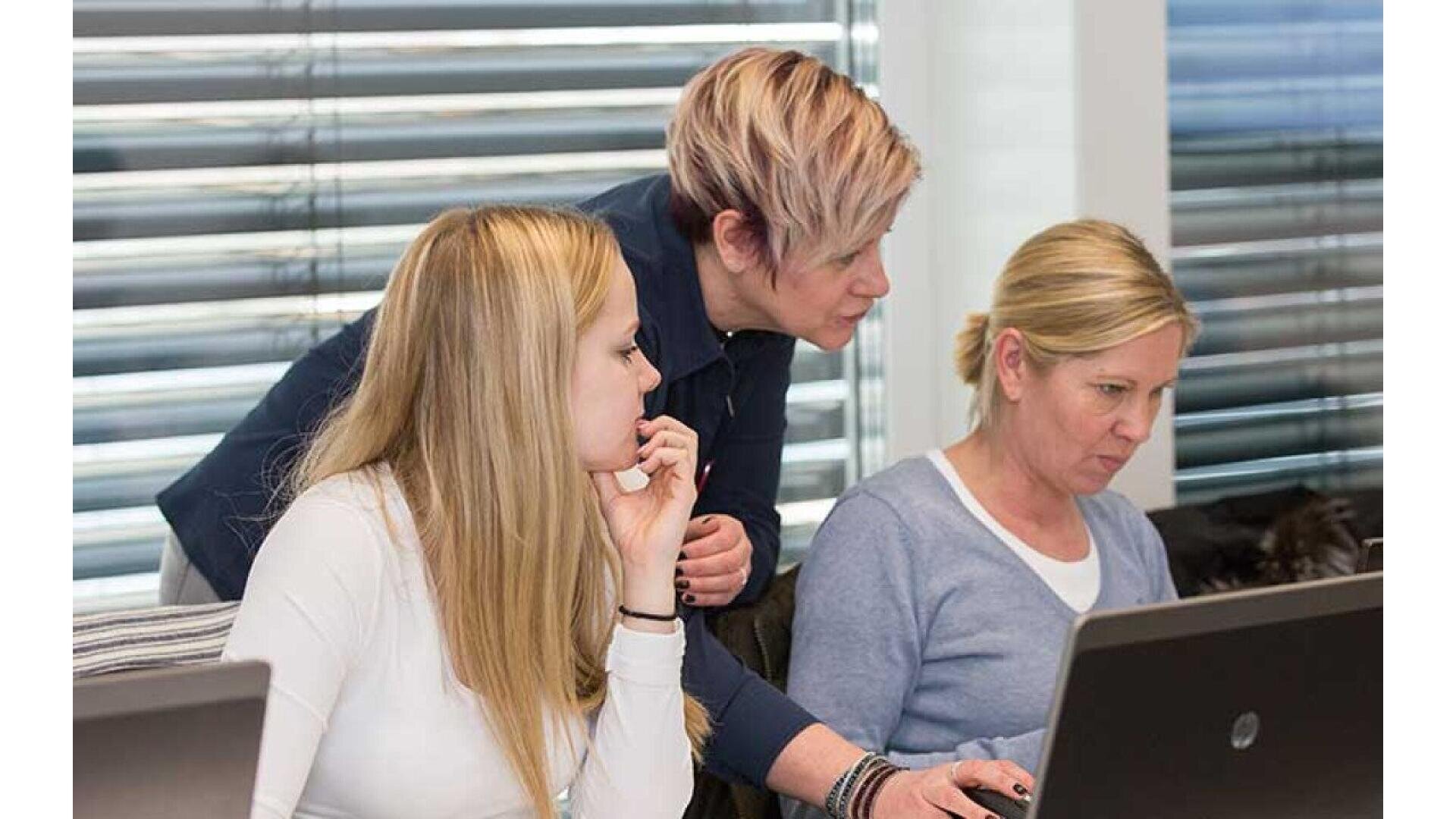 Three women are working together at laptops in an office. One stands and leans over to assist, while the other two sit and focus on their screens. Horizontal blinds cover the window behind them.