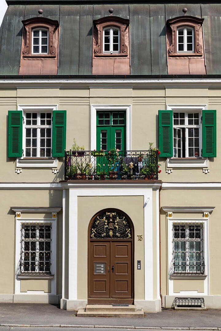 Ein beiges zweistöckiges Gebäude mit grünen Fensterläden, kunstvollen Schmiedearbeiten an Balkon und Fenstern im Erdgeschoss sowie einer braunen Bogentür unter einem mit Topfpflanzen gefüllten Balkon. Auf dem Dach befinden sich drei Dachgauben.
