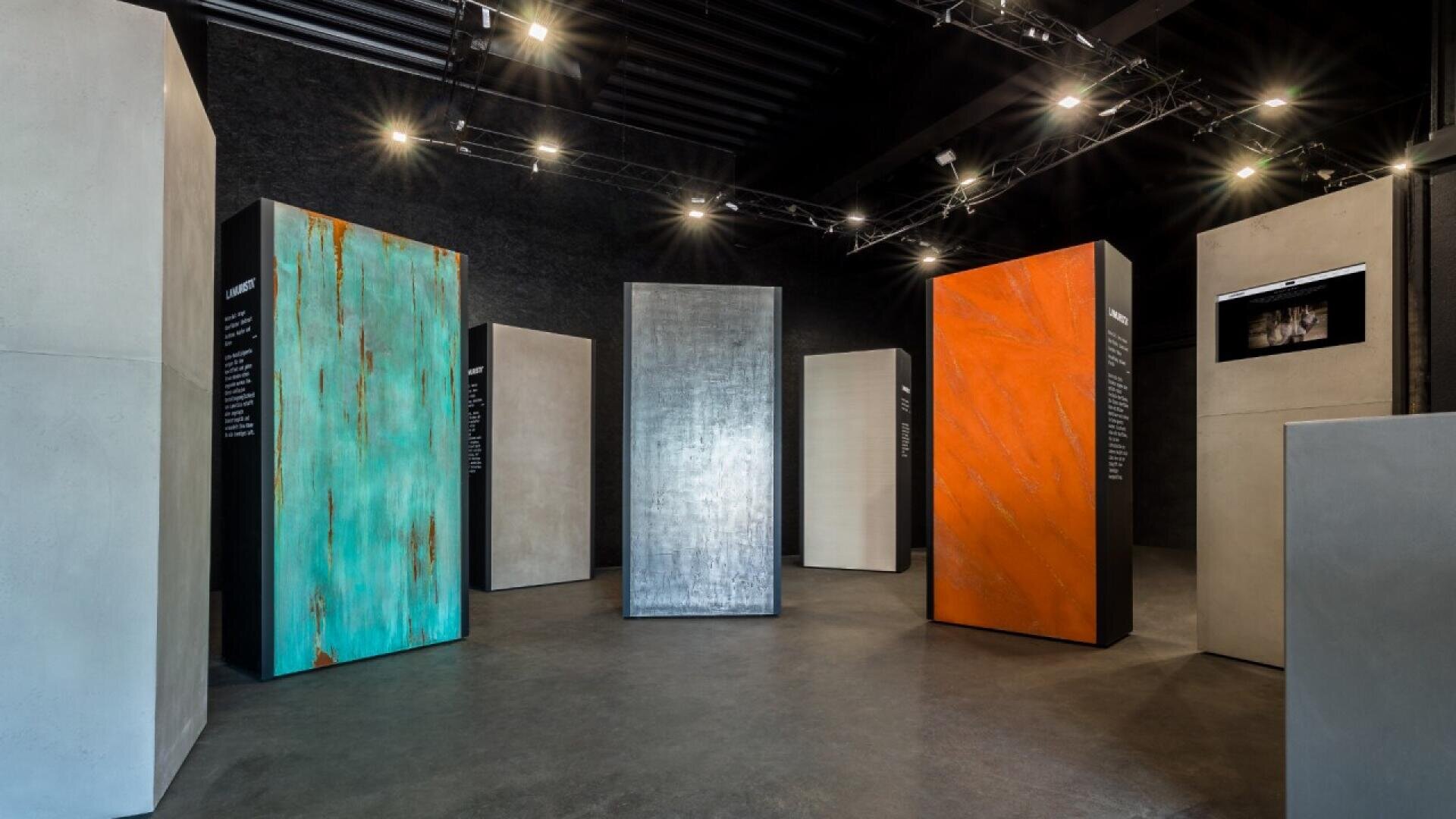 An exhibition room displays several tall, rectangular panels in various colors and textures, including turquoise, metallic silver, and rust orange, arranged in a circle under modern ceiling lights.