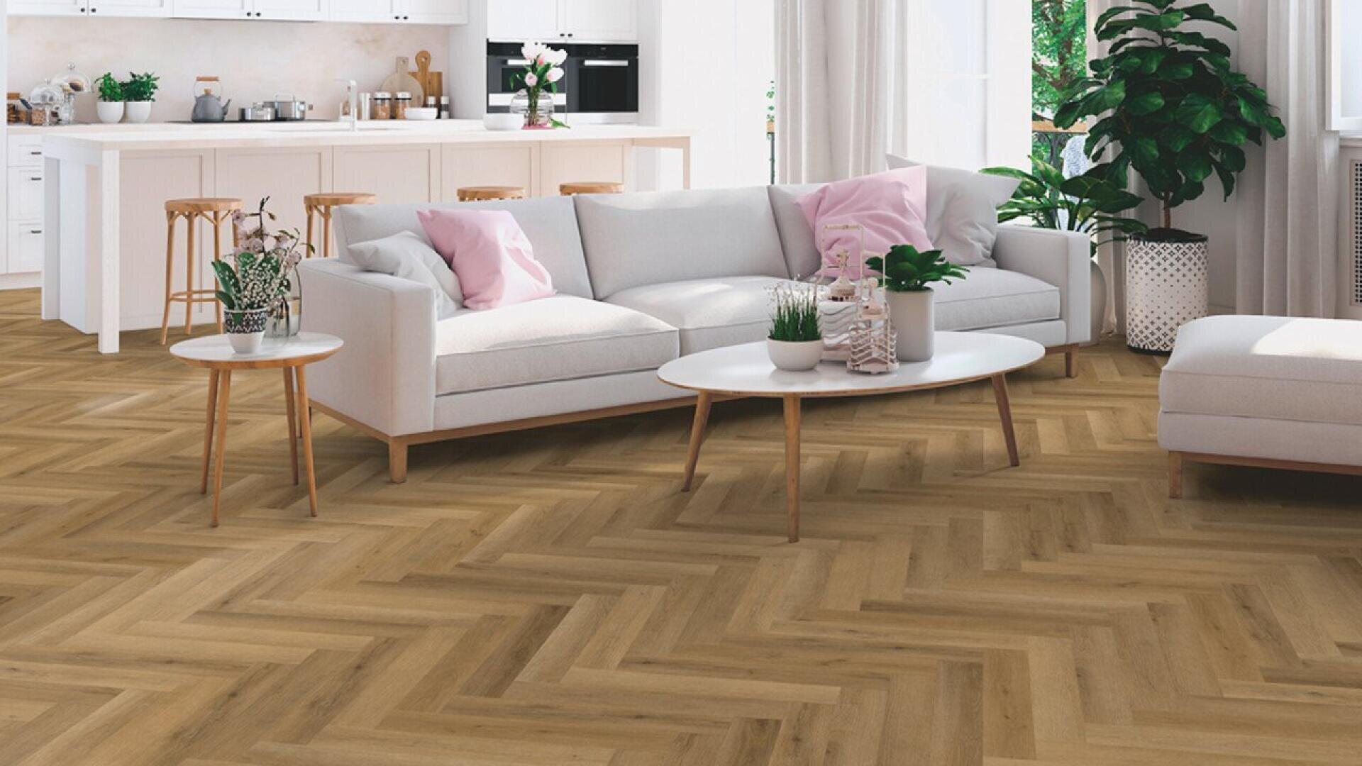 A bright living room with herringbone wood flooring, a white sectional sofa with pink and gray cushions, a round coffee table, and an open kitchen in the background with barstools and white cabinets.