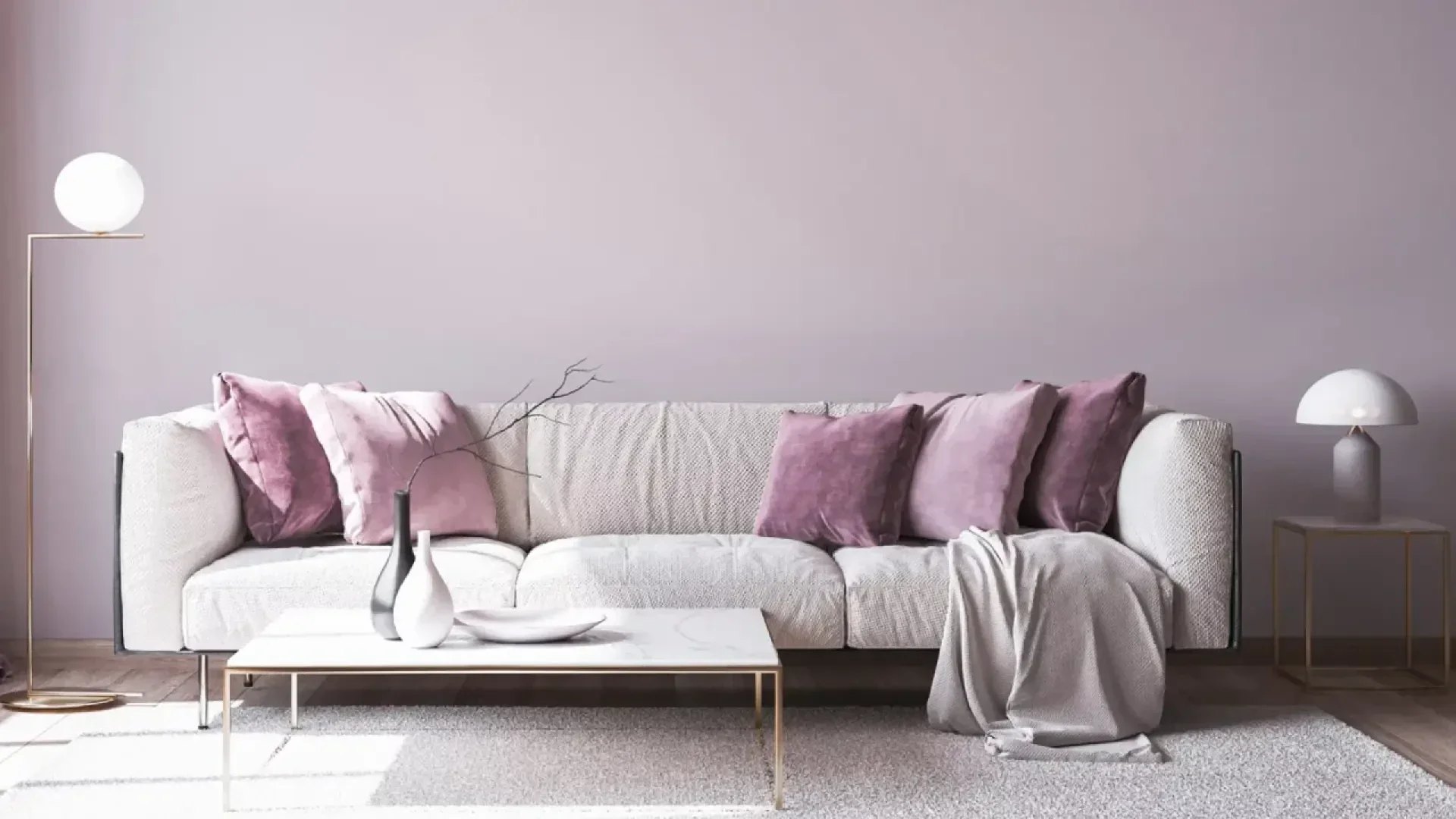A modern living room with a light gray sofa decorated with pink and mauve throw pillows and a gray blanket, a white coffee table with vases, and minimalistic lamps on both sides against a pale lilac wall.
