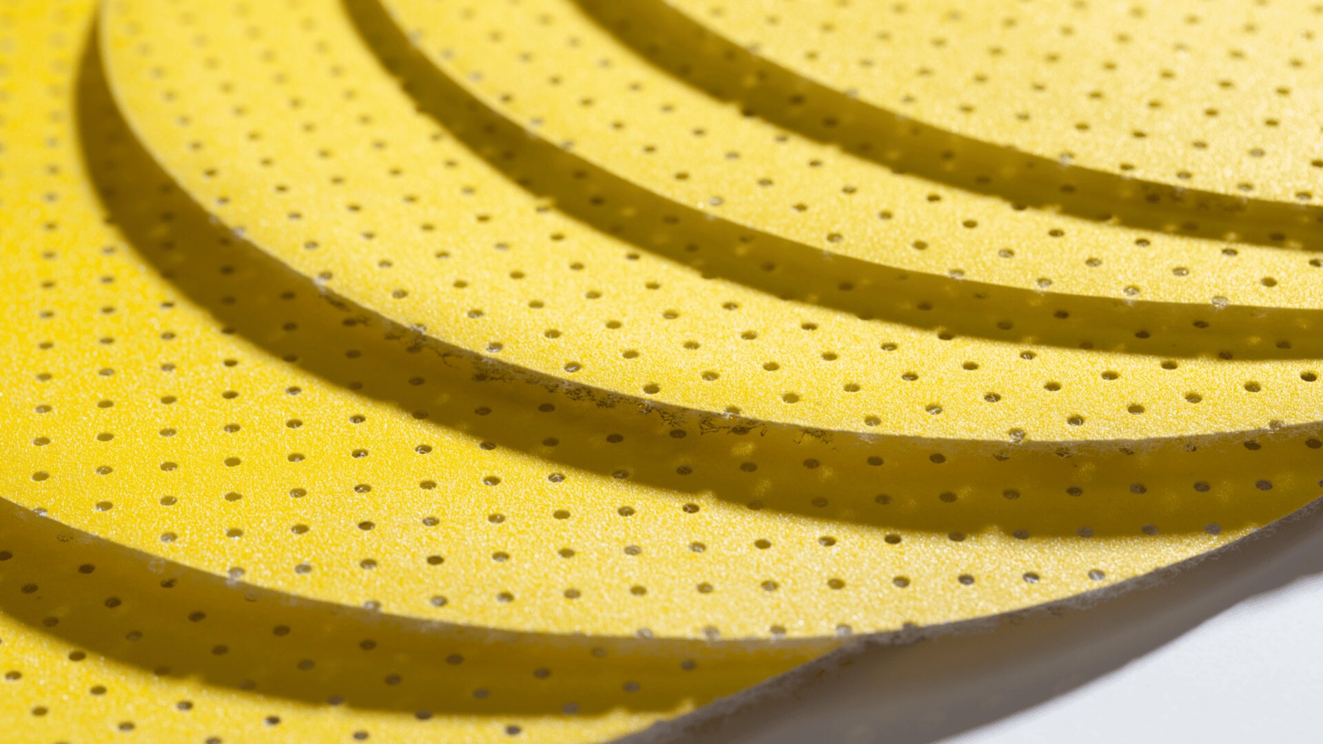 Nahaufnahme mehrerer überlappender, kreisförmiger, gelber Schaumstoffplatten mit kleinen, gleichmäßig verteilten Löchern, die ein geschichtetes und strukturiertes Muster erzeugen.