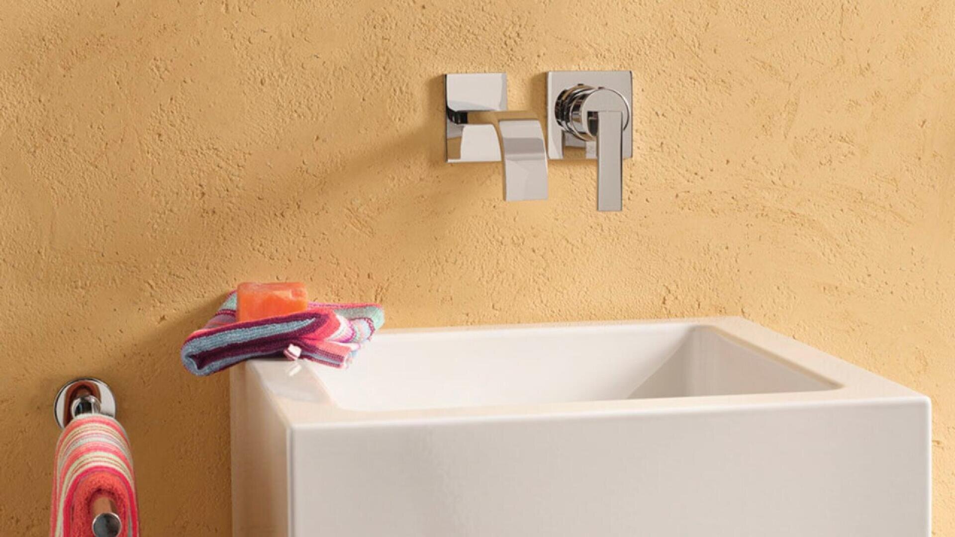 A modern white sink with a chrome wall-mounted faucet is set against a textured beige wall. A striped towel and an orange soap bar rest on the sink’s edge, with another towel hanging nearby.