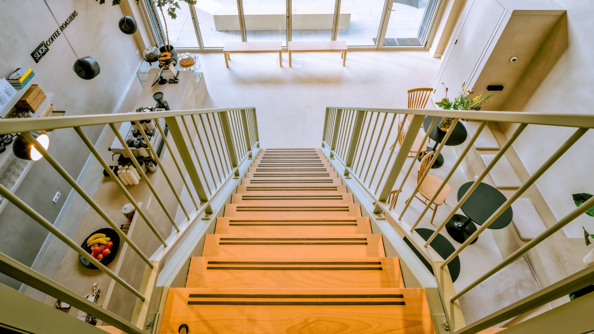 Blick vom oberen Ende einer Holztreppe mit Metallgeländer hinunter in einen modernen, hellen Wohnraum mit großen Fenstern, minimalistischen Möbeln und einem kleinen Essbereich.