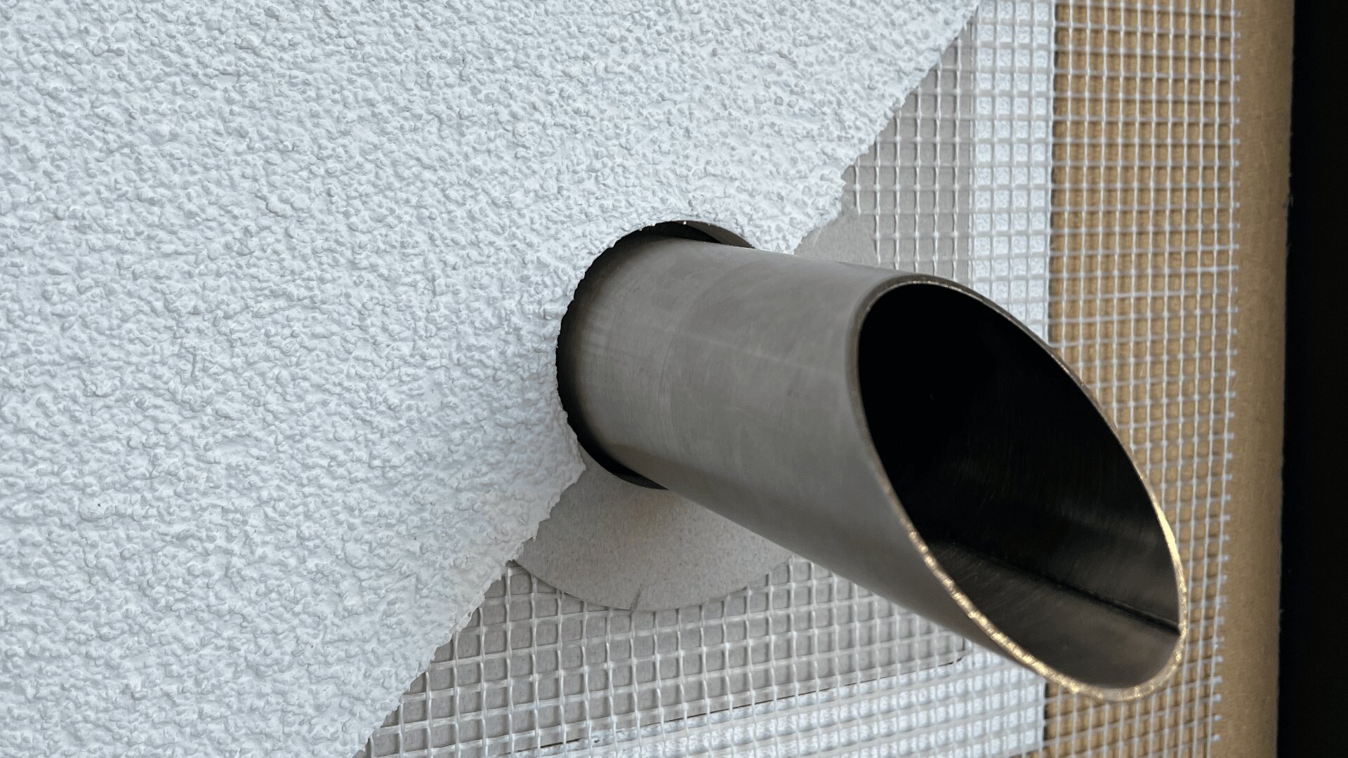 Eine Nahaufnahme eines Metallrohrs, das schräg aus einer strukturierten weißen Wand herausragt, wobei um die Rohröffnung herum ein kreisförmiger Ausschnitt und eine Netzverstärkung sichtbar sind.