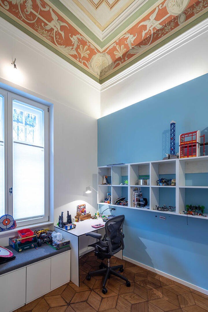 Ein helles, modernes Arbeitszimmer mit einer hohen, verzierten Decke, einer blauen Akzentwand, weißen Regalen mit Spielzeug und Modellen, einem Schreibtisch mit Stuhl und einem Fenster mit einer gepolsterten Bank darunter.