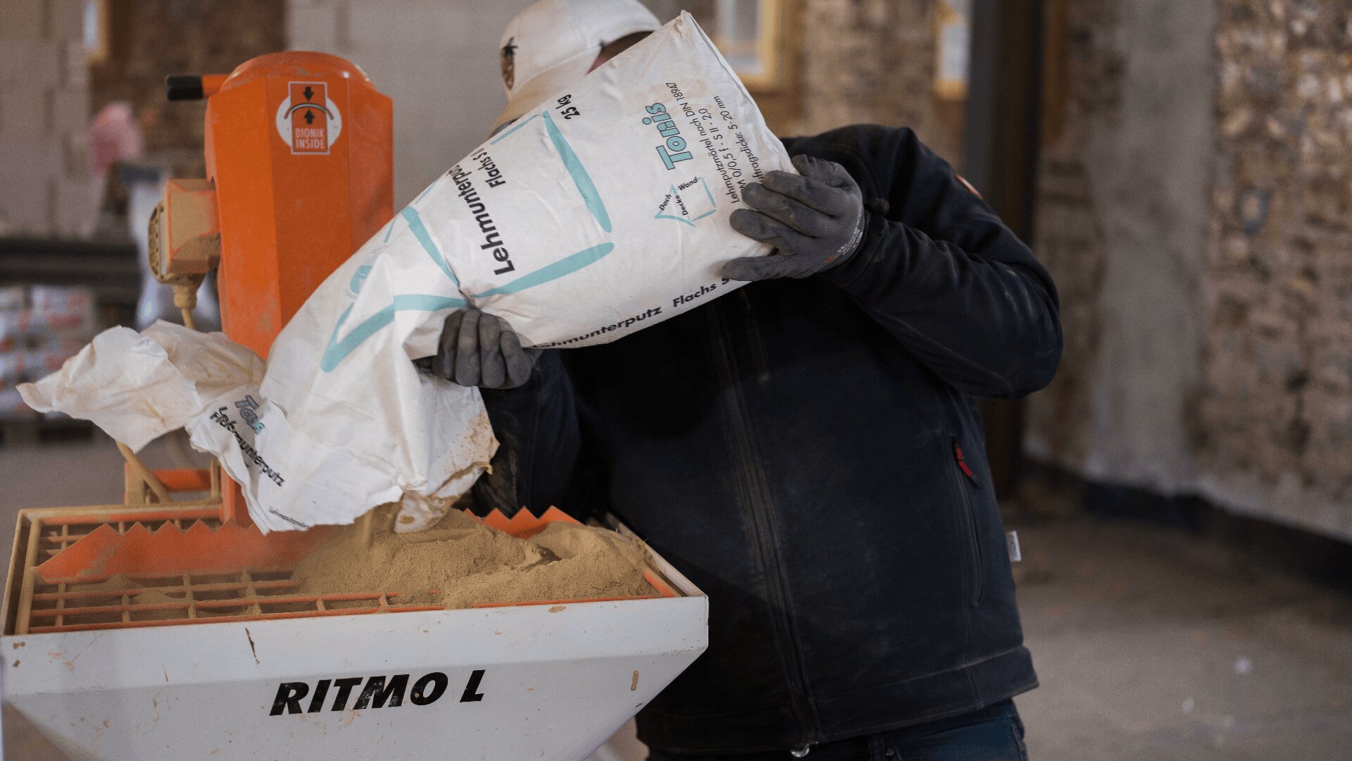 Ein Bauarbeiter mit Schutzhelm schüttet auf einer Baustelle einen Sack mit Material mit der Aufschrift „Lemmenpulver“ in eine Mischmaschine mit der Aufschrift „RITMO L“.