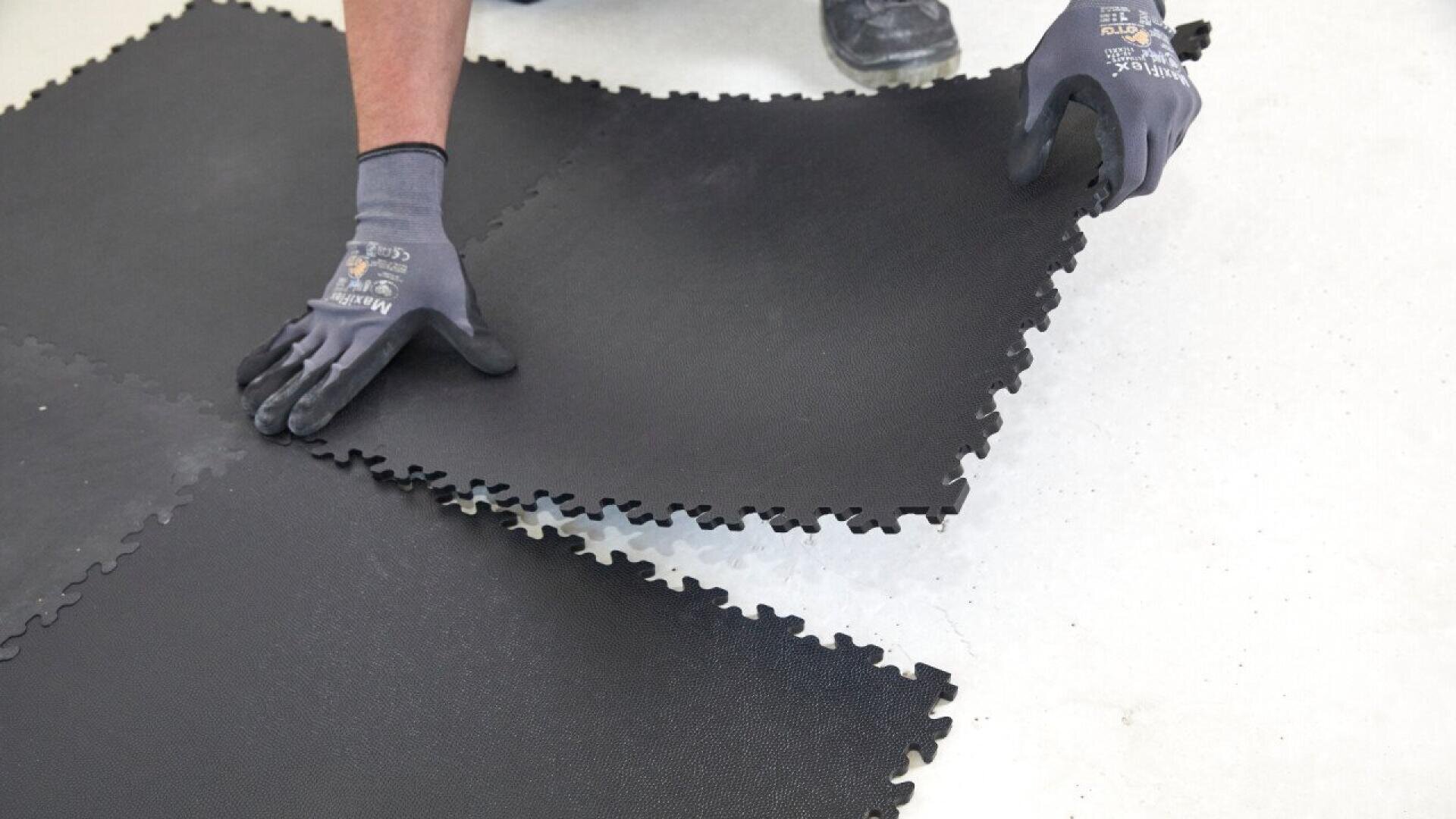 A person wearing gloves fits interlocking black rubber floor tiles together on a light-colored surface.
