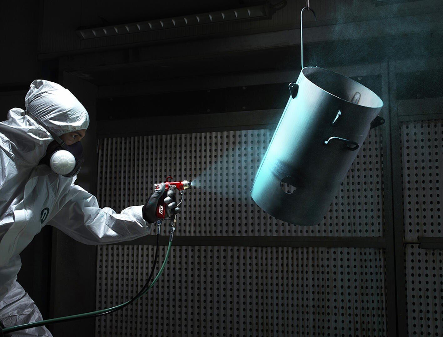 Eine Person in Schutzkleidung und Atemschutzmaske sprüht einen großen Metallbehälter, der in einer Werkstatt hängt, mit einer an einen Schlauch angeschlossenen Spritzpistole. Im Hintergrund sind Lochbleche und Industriebeleuchtung zu sehen.