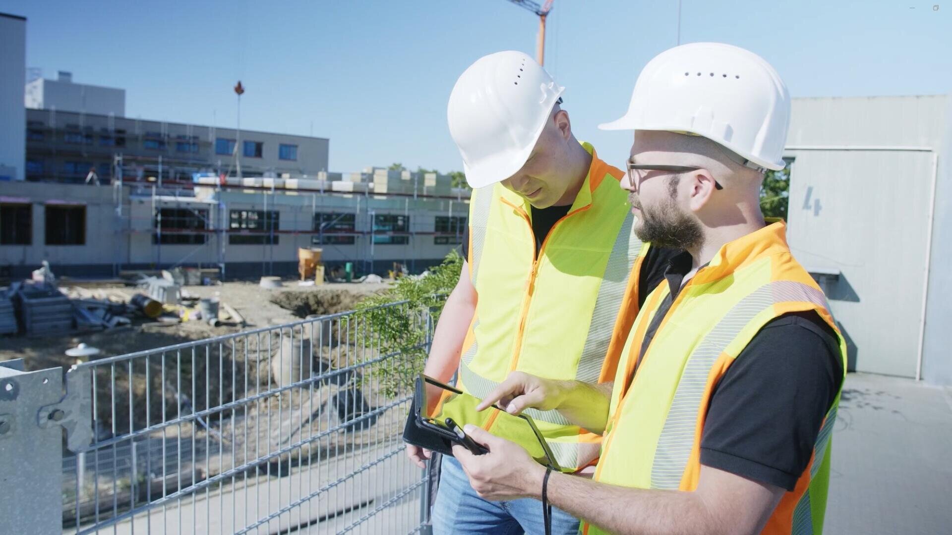 Zwei Bauarbeiter in Sicherheitswesten und weißen Helmen stehen draußen auf einer Baustelle und betrachten ein Gerät, möglicherweise eine Drohnensteuerung, während im Hintergrund ein im Bau befindliches Gebäude zu sehen ist.