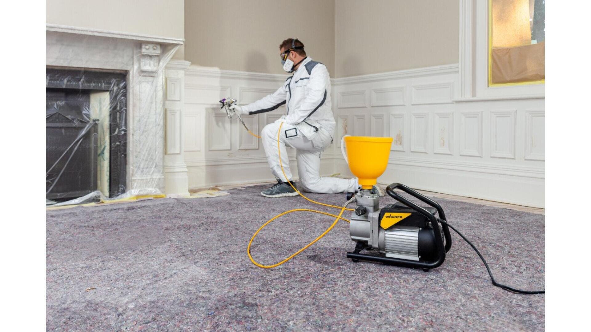 A person wearing protective gear uses a paint sprayer to paint white wainscoting in a room with covered floors and masked windows. A yellow and black paint sprayer machine sits on the floor in the foreground.