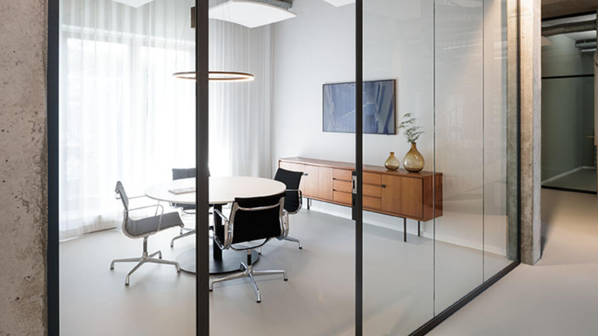 Ein moderner Büro-Besprechungsraum mit einem runden weißen Tisch, vier schwarzen Stühlen, einem hölzernen Sideboard mit Vasen, einem gerahmten Bild an der Wand und großen Fenstern mit transparenten Vorhängen. Der Raum ist durch Glaswände abgetrennt.