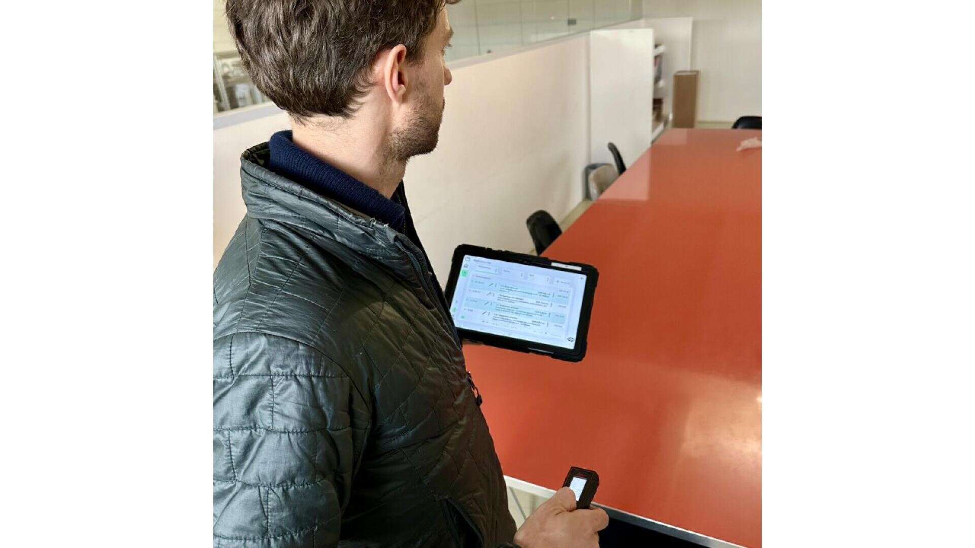 A man in a gray jacket holds a tablet displaying a digital form while standing near a long red table in a modern office or meeting room.