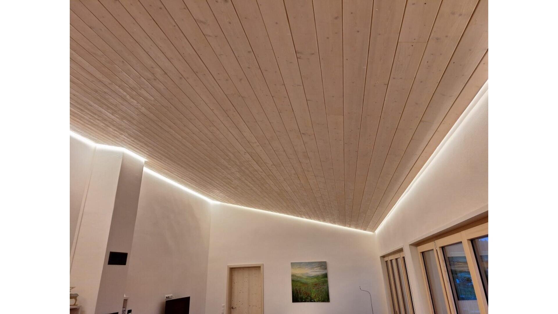 A modern room with a slanted, light wooden ceiling, illuminated by concealed strip lighting along the edges. The walls are white, with a door, a landscape painting, and large windows on one side.