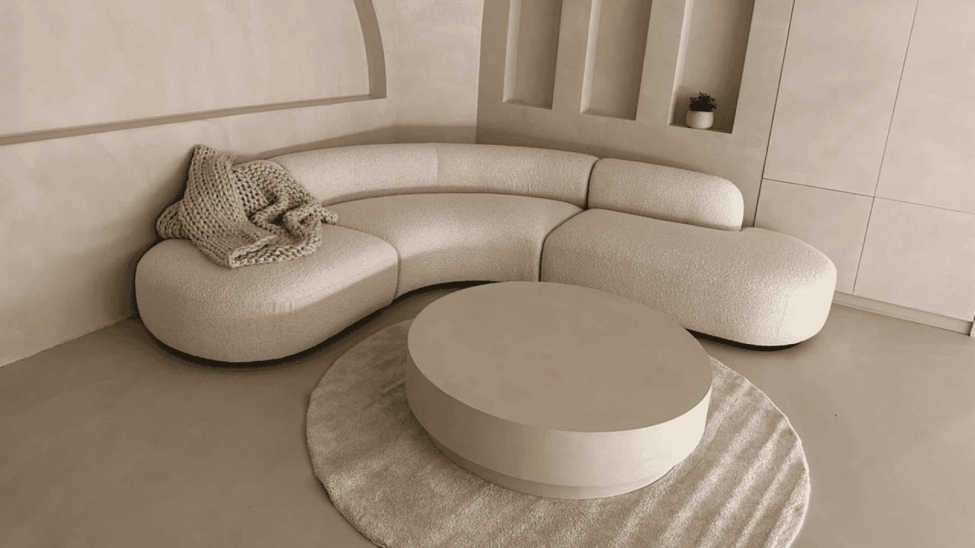A modern living room with a curved cream sofa, a chunky knit blanket, a round coffee table, and a circular rug. The walls feature geometric cutouts and a small potted plant. The decor is minimalist and neutral-toned.