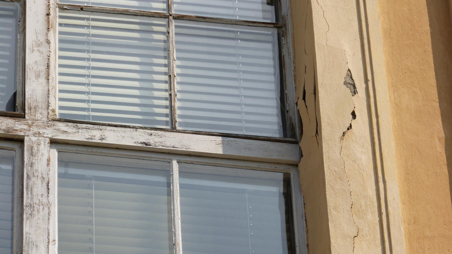 Nahaufnahme eines Altbaufensters mit weißen Jalousien dahinter. Die gelbe Außenwand ist rissig und blättert ab, der Fensterrahmen wirkt verwittert und abgenutzt. Sonnenlicht wirft Schatten auf die Oberfläche.