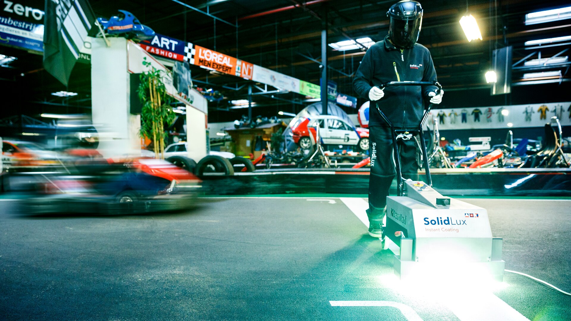 Eine Person in Schutzkleidung bedient eine SolidLux-Maschine, die eine leuchtend weiße Linie auf eine Indoor-Gokartbahn malt, während im Hintergrund ein Gokart vorbeifährt.