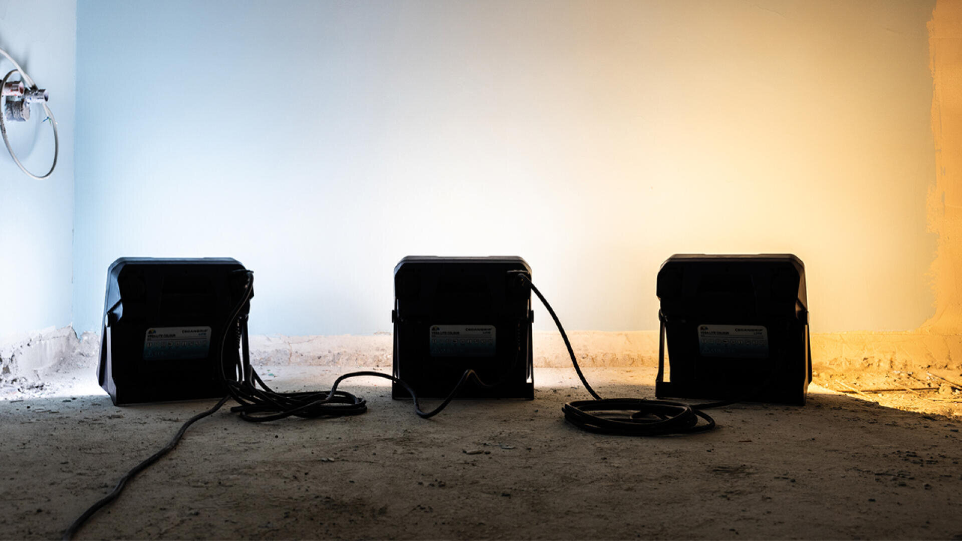 Three black portable devices with cables are placed on a rough, unfinished floor against a wall that is lit with both cool white and warm yellow lighting. An exposed electrical outlet is visible on the left.