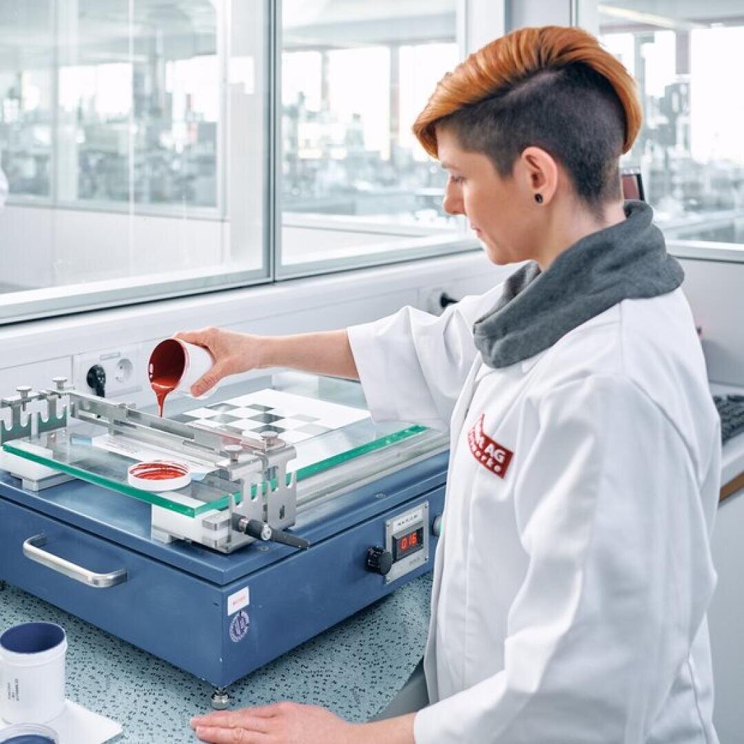 Eine Person im Laborkittel gießt in einer Laborumgebung eine rote Flüssigkeit auf eine Glasplatte. Dabei verwendet sie wissenschaftliche Geräte auf einer Arbeitsplatte, daneben stehen Becher.