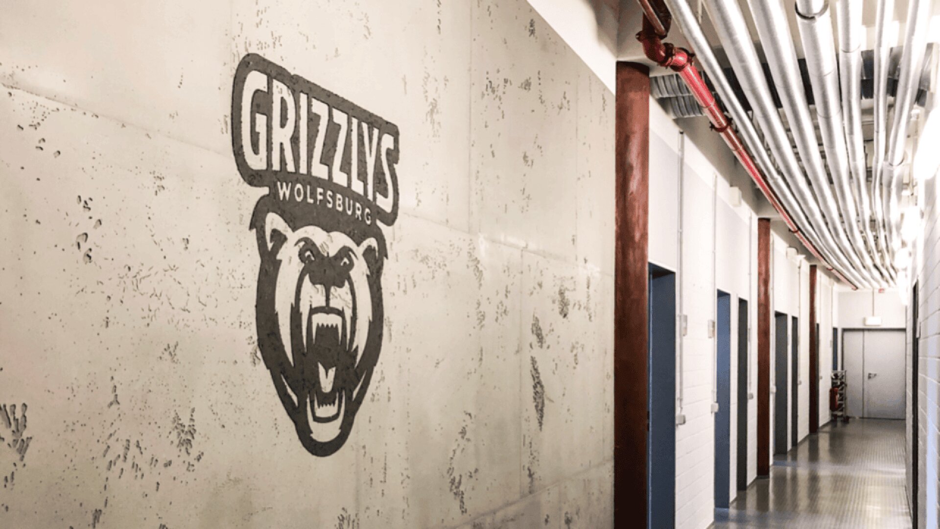 A hallway with industrial pipes on the ceiling and several doors. The wall features a large Grizzlys Wolfsburg logo with a roaring bear graphic.