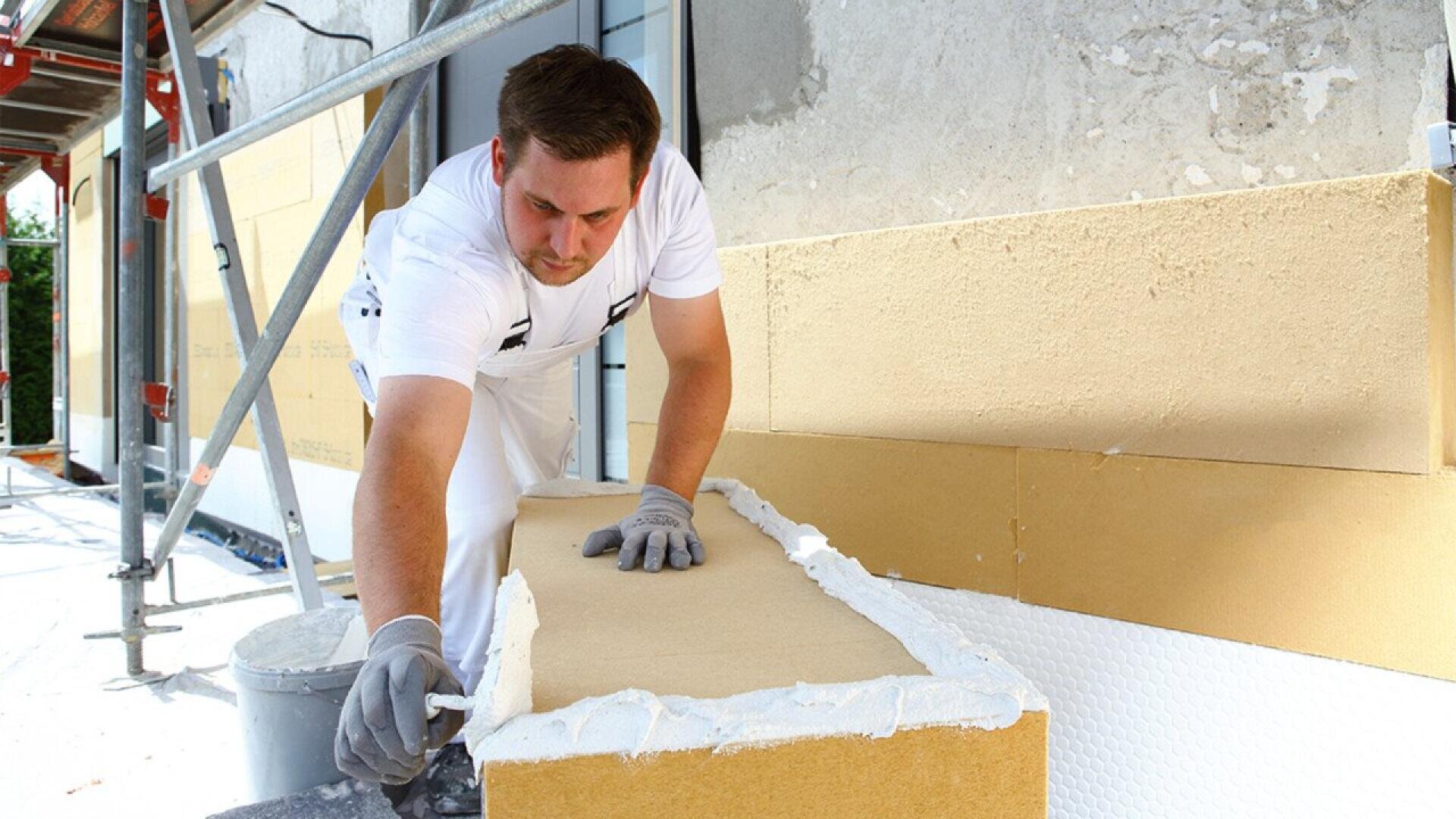 A construction worker in white overalls applies adhesive to a large yellow insulation panel on the exterior wall of a building under renovation. Scaffolding and tools are visible nearby.