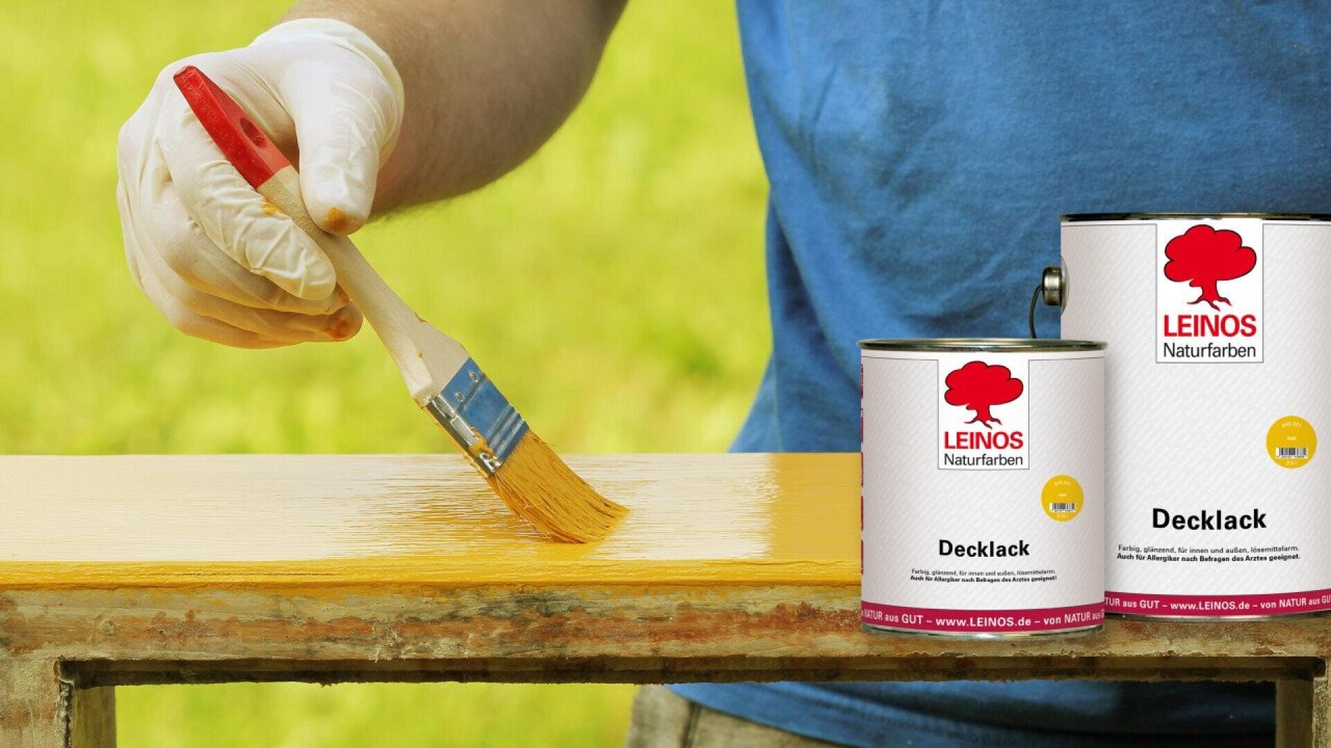 A person wearing a white glove applies yellow paint to a wooden board with a brush. Two cans of LEINOS Decklack paint are shown in the foreground with a blurred green background.