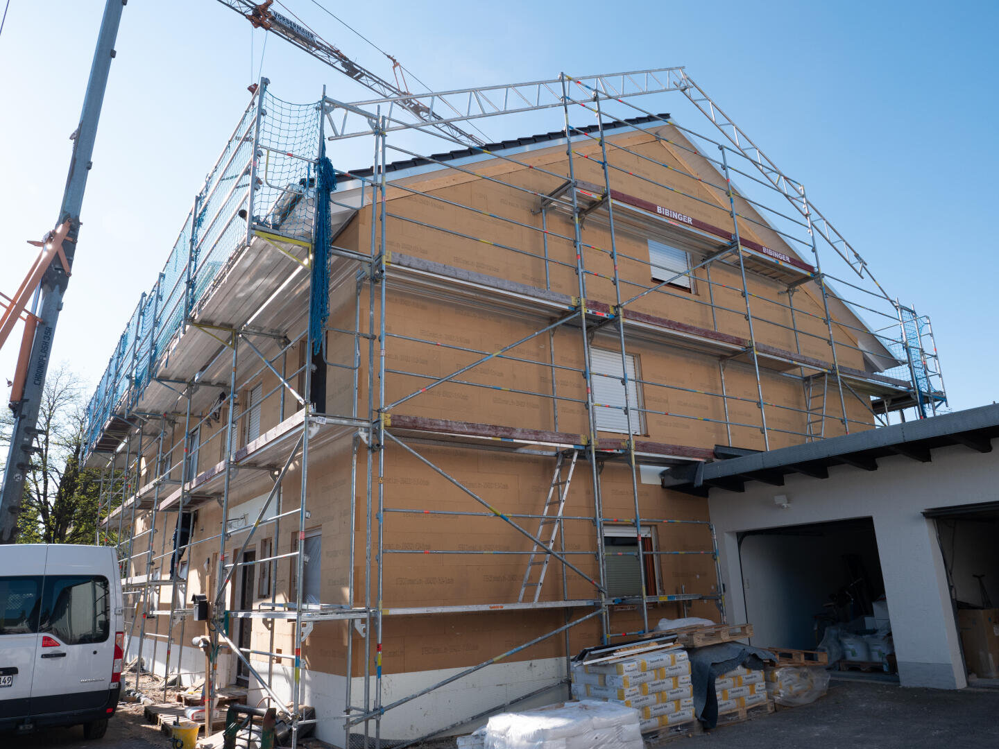 Ein zweistöckiges Haus im Bau, umgeben von einem Gerüst; in der Nähe stapeln sich Baumaterialien, links parkt ein weißer Lieferwagen. Der Himmel ist klar und blau.