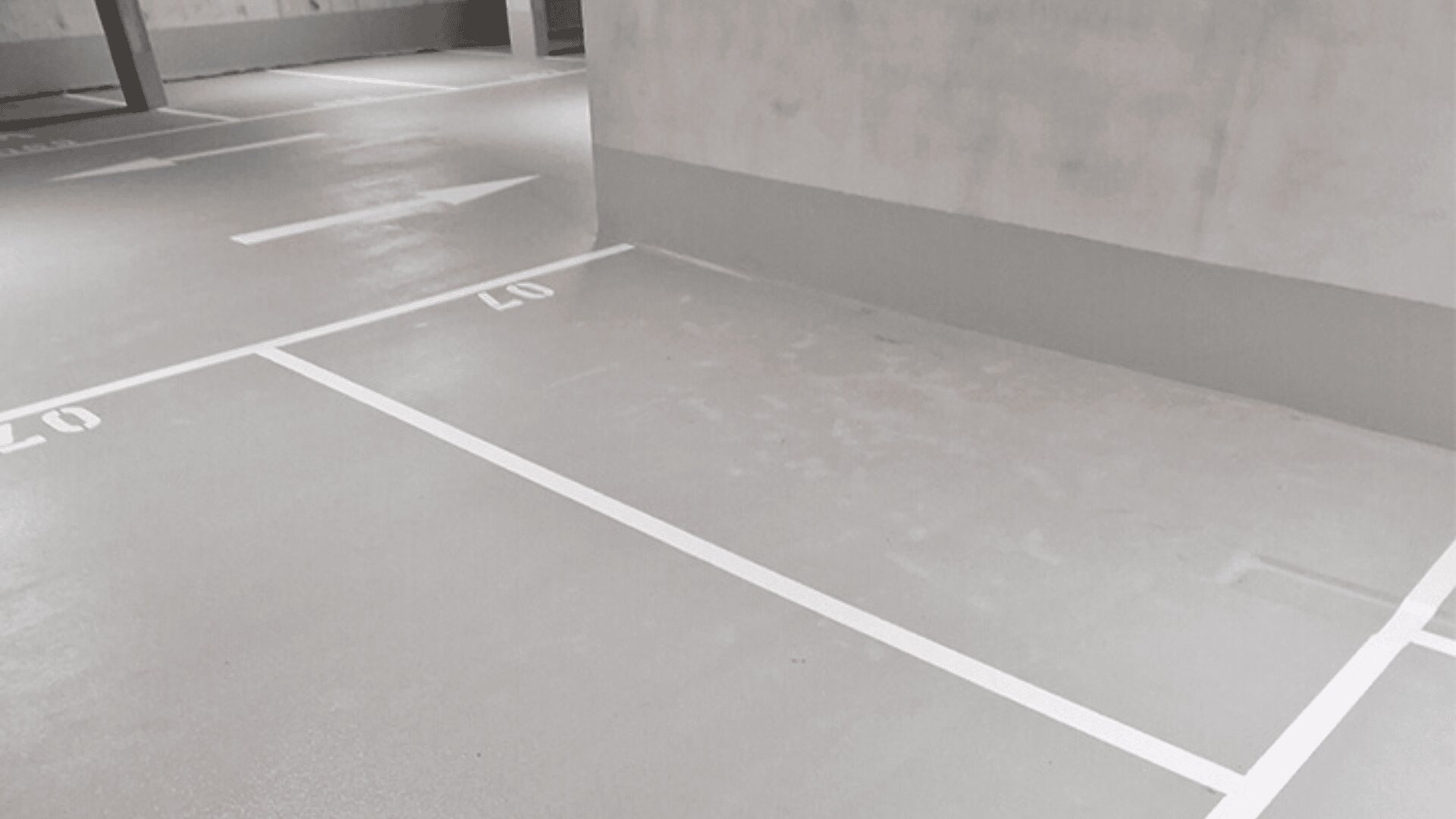 Ein leeres Parkhaus mit deutlich markierten weißen Parkplätzen und Pfeilen auf einem sauberen, grauen Boden. Der sichtbare Parkplatz trägt die Nummer 07, und eine Betonwand versperrt teilweise die Sicht nach vorne.
