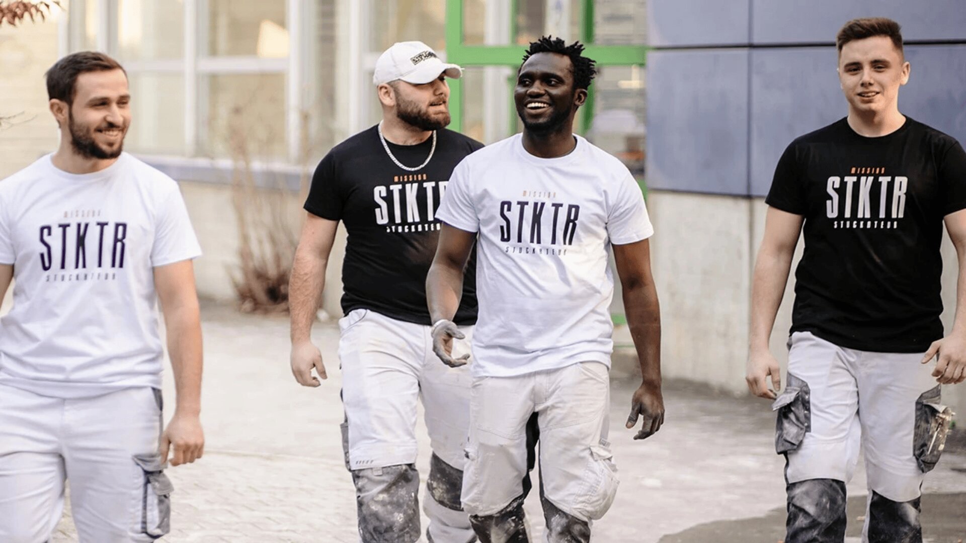 Vier Männer in passenden STKTR-T-Shirts und farbbeklecksten Arbeitshosen gehen gemeinsam vor einem Gebäude spazieren, lächeln und unterhalten sich, was auf Kameradschaft und Teamwork hindeutet.