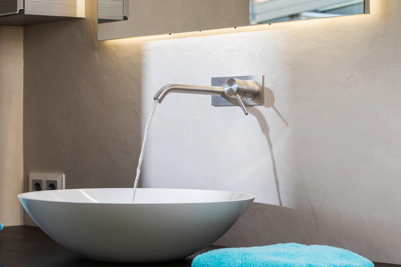 Ein an der Wand montierter Wasserhahn leitet Wasser in ein modernes weißes Aufsatzwaschbecken unter einem Spiegel, mit sanfter Beleuchtung und türkisfarbenen Handtüchern auf einer dunklen Arbeitsplatte in einem minimalistischen Badezimmerambiente.