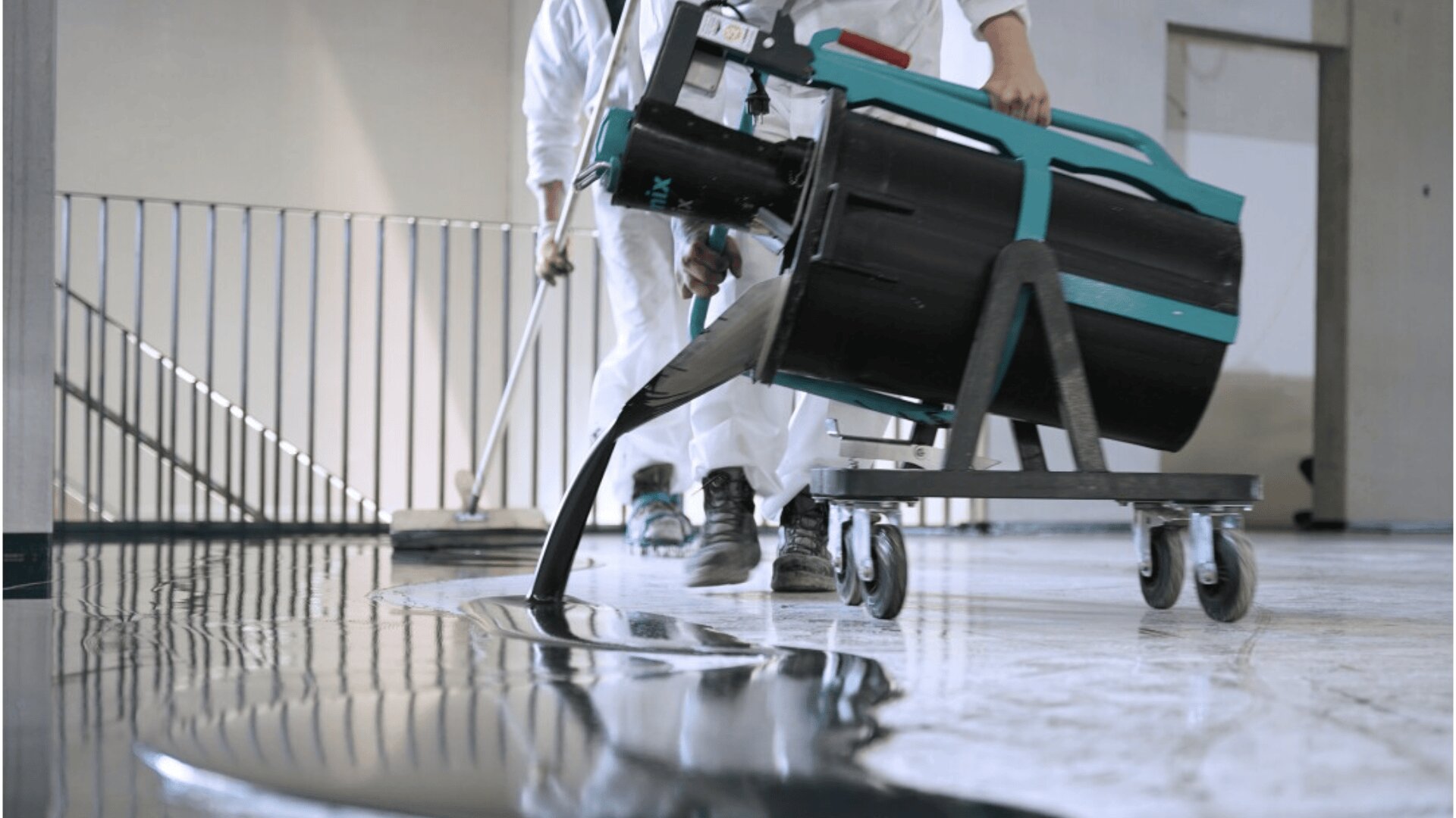 Two workers in white protective suits pour a dark liquid from a machine onto a floor, possibly applying an epoxy or resin coating, with one person using a tool to spread the material.